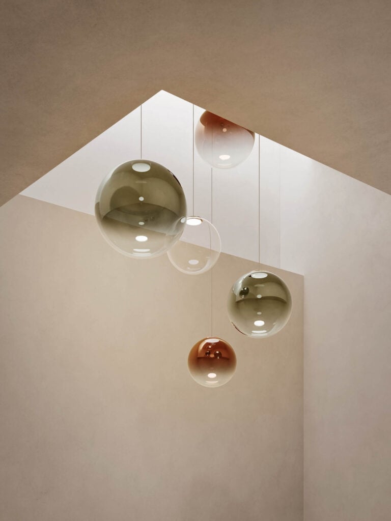 Four glass pendant lights in various sizes and colors hang from the ceiling of a minimalist room with neutral-toned walls and an open skylight above.