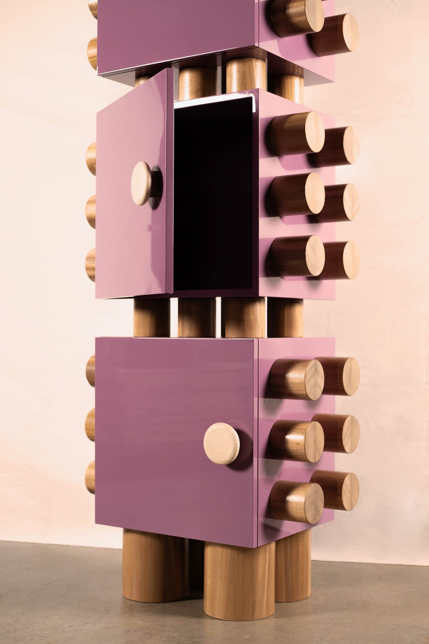 Tall, geometric cabinet with glossy purple panels, cylindrical wooden handles, and legs. One door is open, revealing a dark interior. The design is bold and modern.