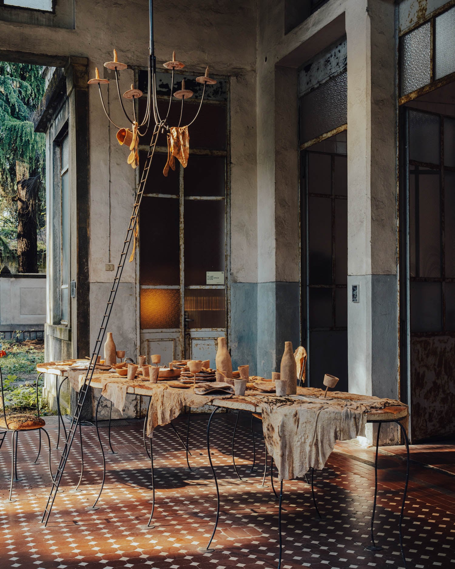An indoor table set with rustic, clay pottery and textiles sits beneath an unusual chandelier, in a sunlit, weathered room with tiled floors and large windows.