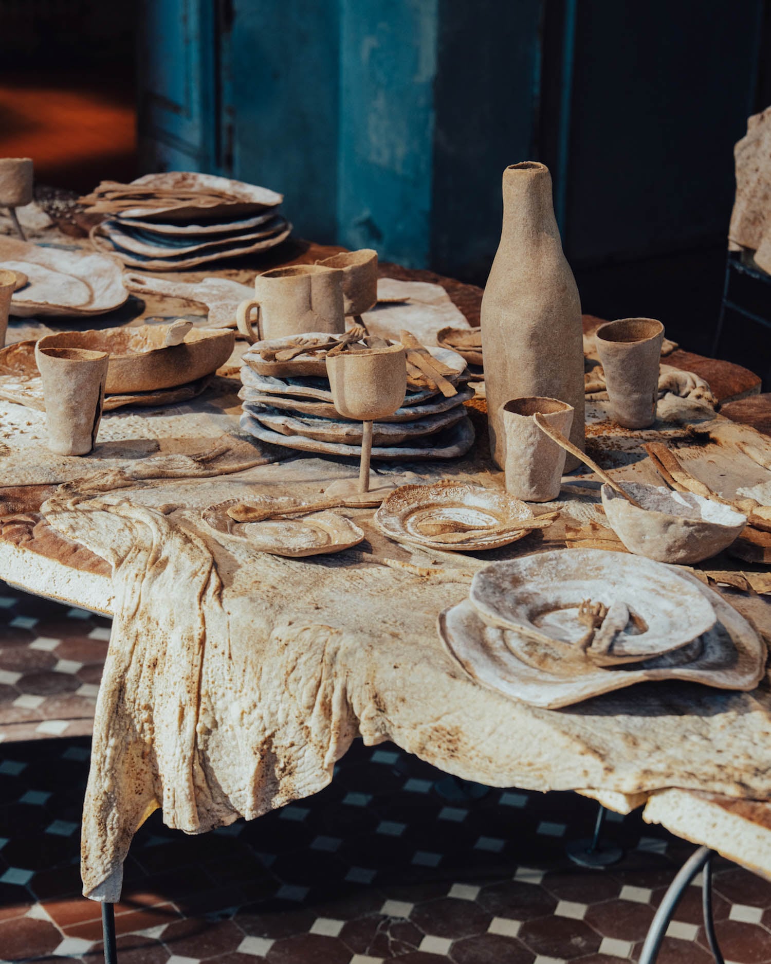 A rustic table set with plates, cups, bowls, and a bottle made from earthy, textured materials, all covered in a dusting of flour on a similarly textured tablecloth.