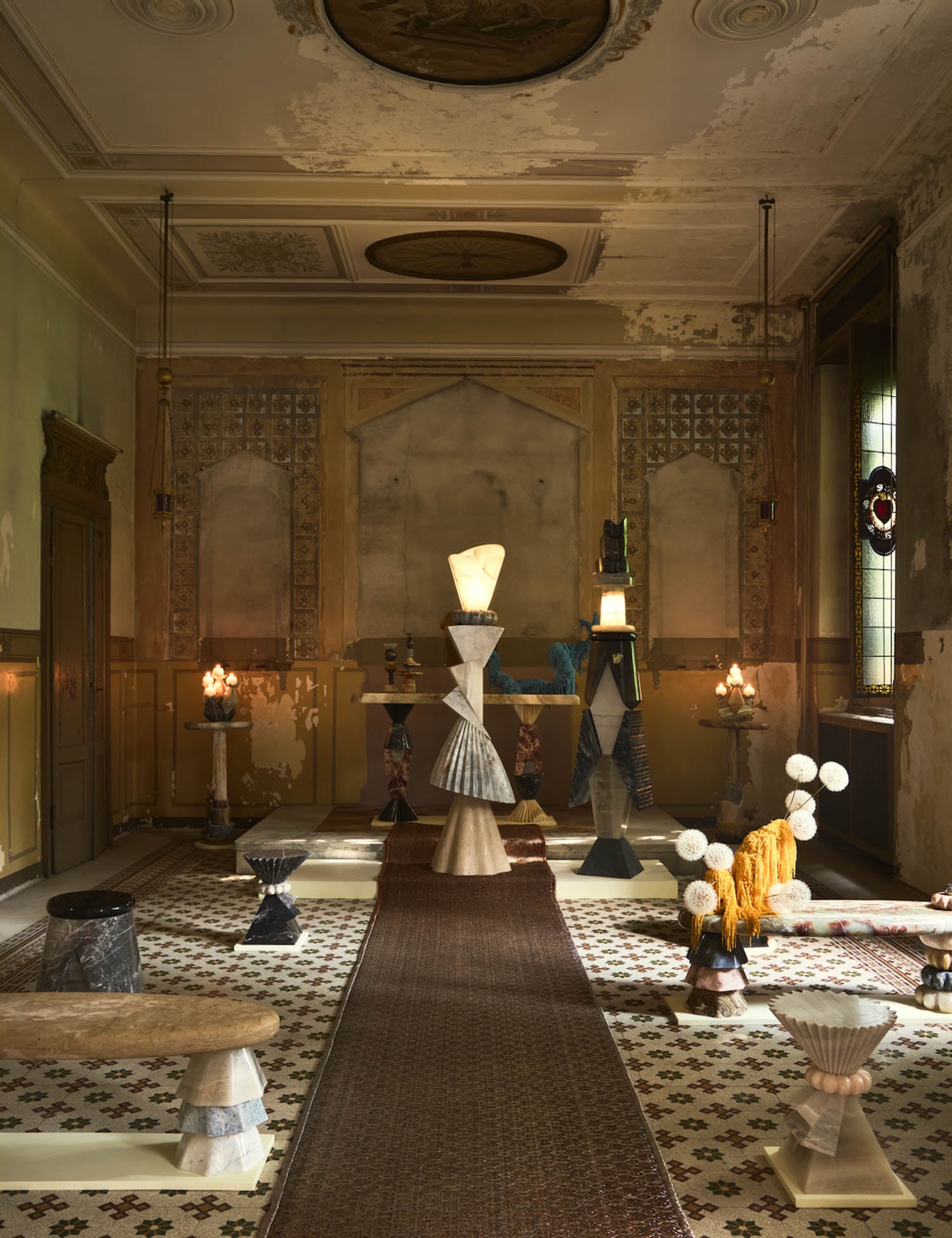 A dimly lit, ornate room features eclectic sculptural furniture and decor arranged on a patterned tile floor, with weathered walls and stained glass windows.