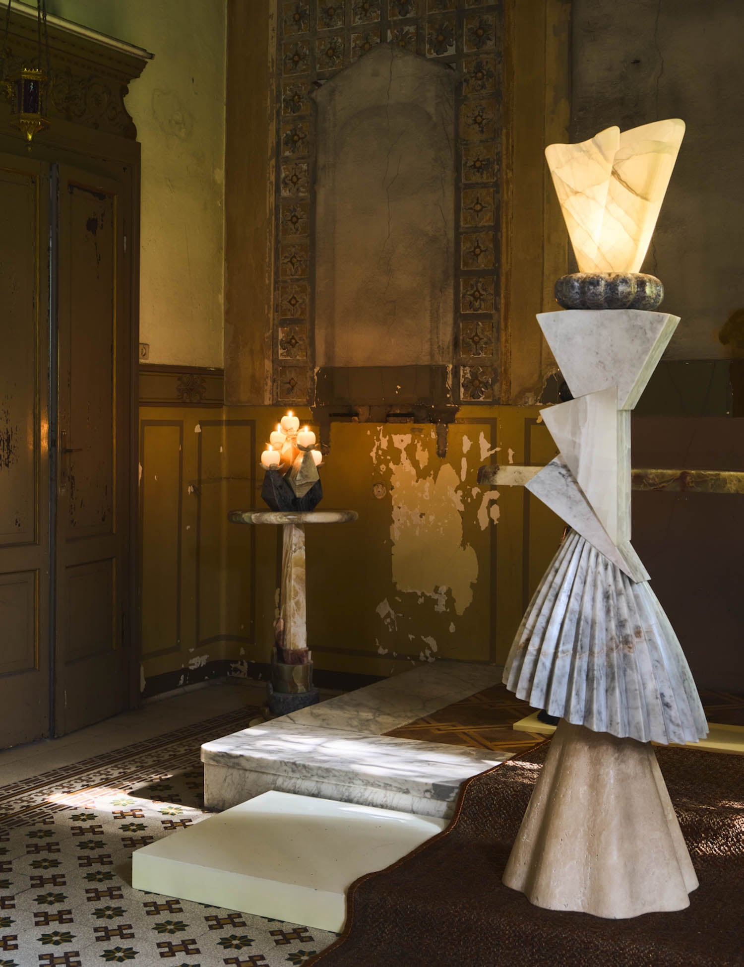 A dimly lit room with worn walls features geometric stone and light sculptures, including a tall illuminated piece in the foreground and a smaller lamp on a round table.