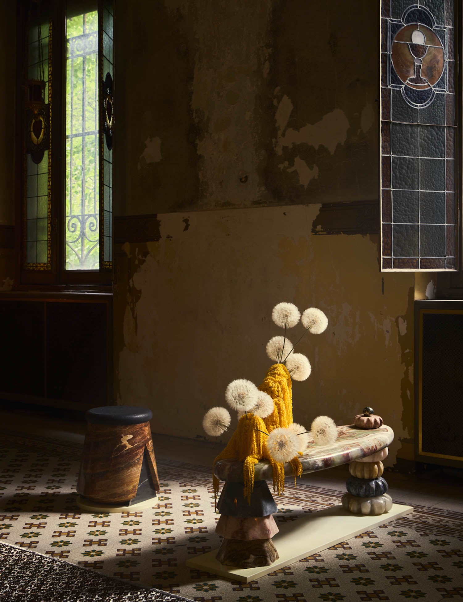 A small, dimly lit room features stained glass windows, a patterned tile floor, a rustic stool, and an abstract table with a sculptural floral arrangement.