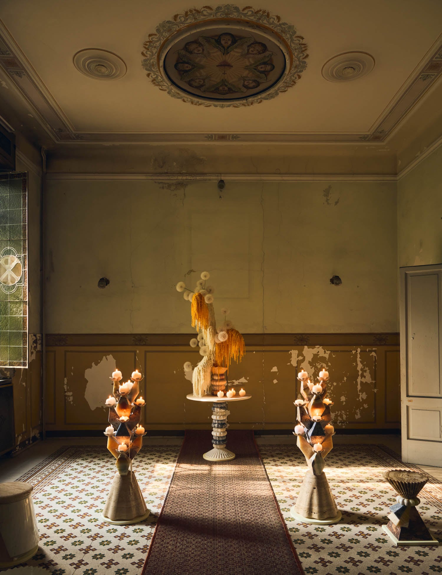 Dimly lit room with peeling paint, ornate ceiling, tiled floor, and three candle arrangements on pedestals surrounding a central table with decorative objects and candles.