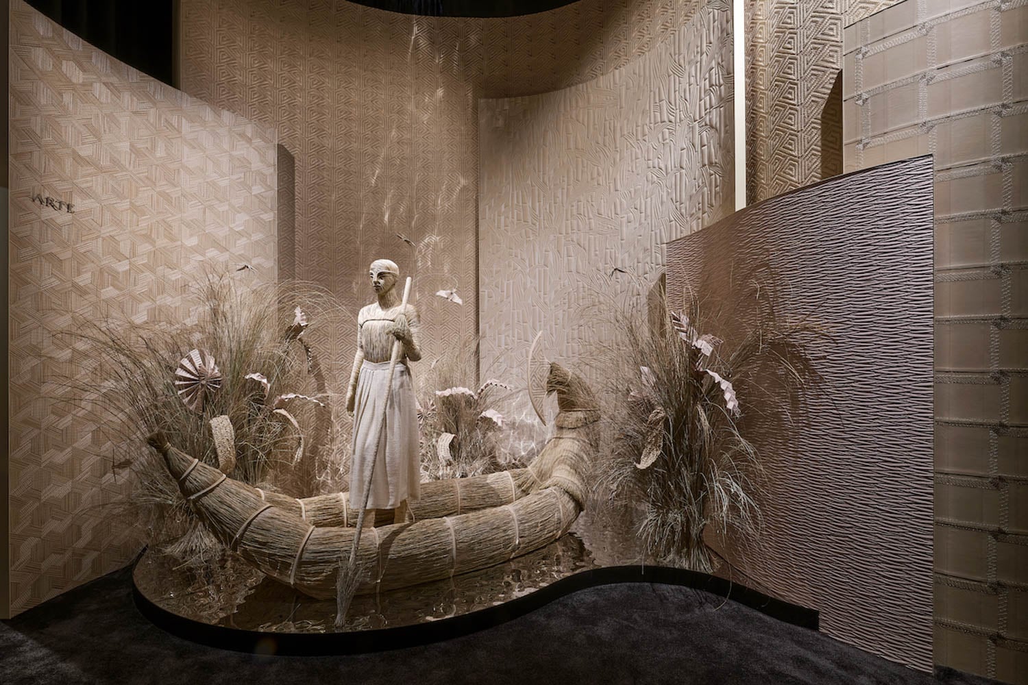 A mannequin in a white dress stands in a woven boat surrounded by dry grasses and textured beige panels in an artfully designed display.