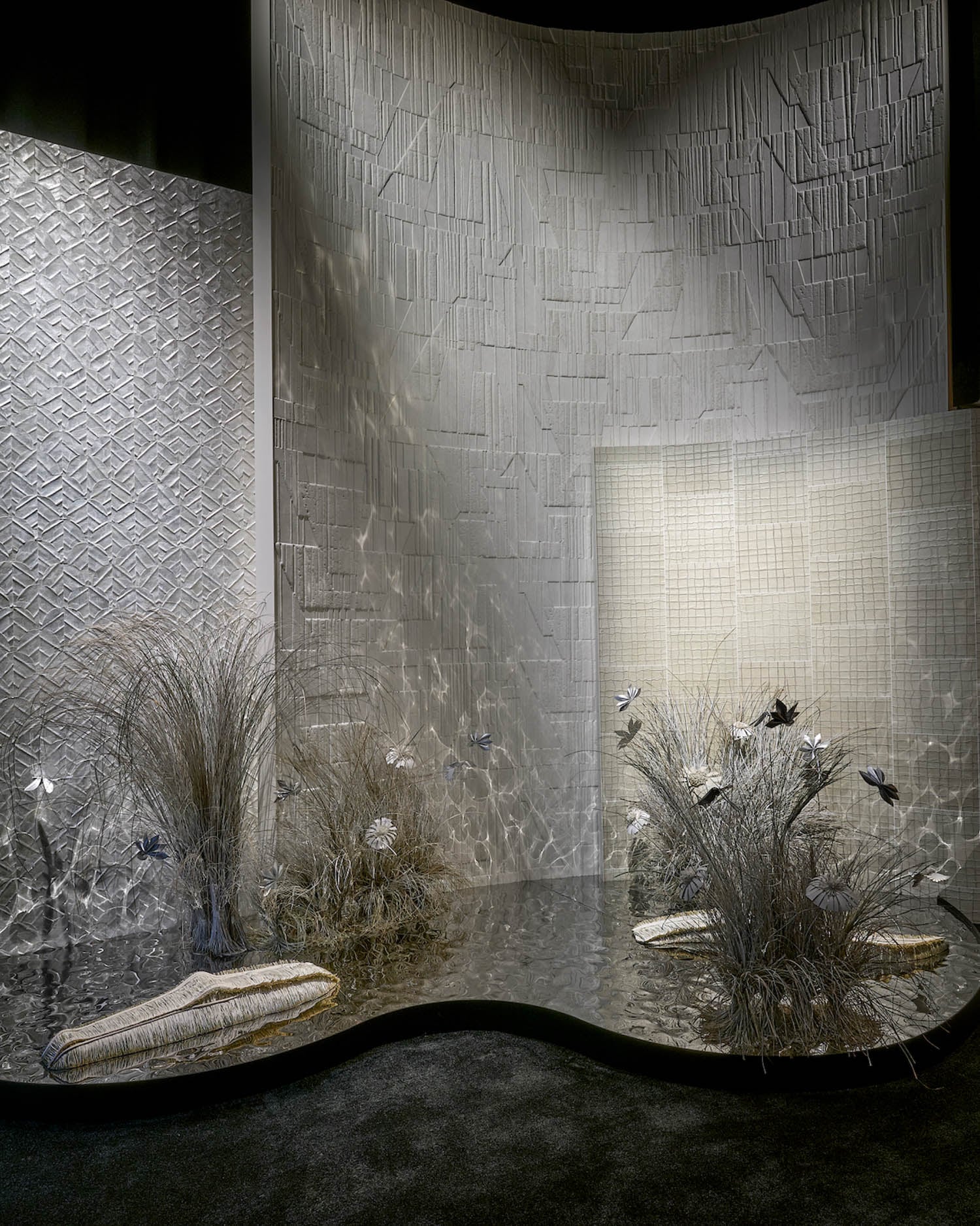 Curved wall panels with textured geometric patterns, metallic flooring, decorative dried grasses, flowers, and two woven cushions arranged in a minimalist, modern display.