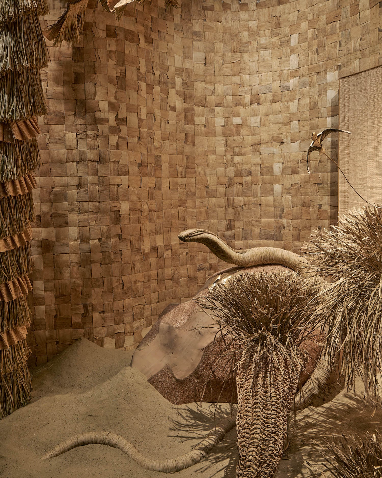 A room with woven straw walls, sand-covered floor, large woven rope sculptures resembling snakes, and dry plant-like structures creating a desert-like scene.
