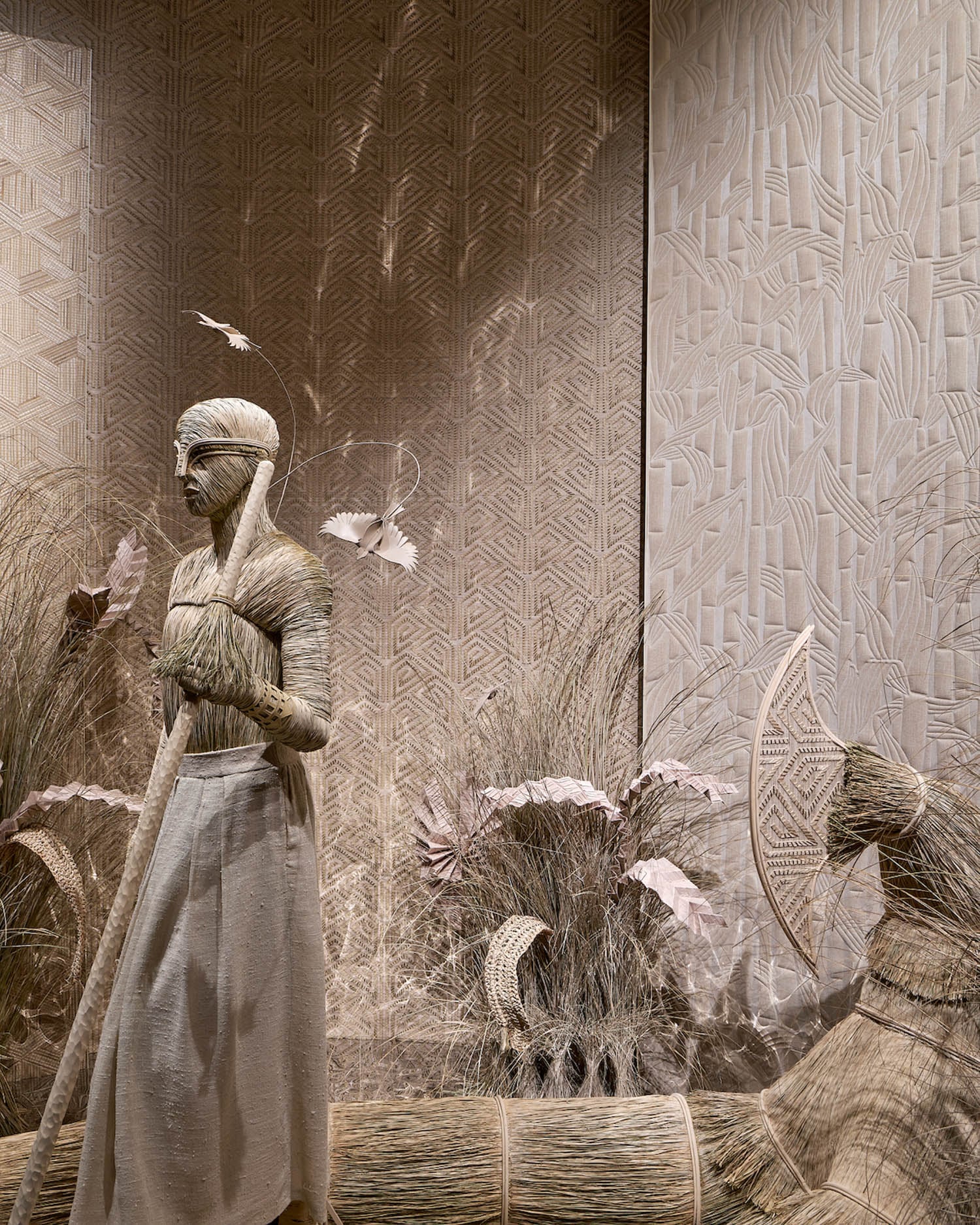 A woven sculpture of a person holding a staff stands beside a decorative boat and grasses, all crafted from natural fibers, against a textured, patterned wall.