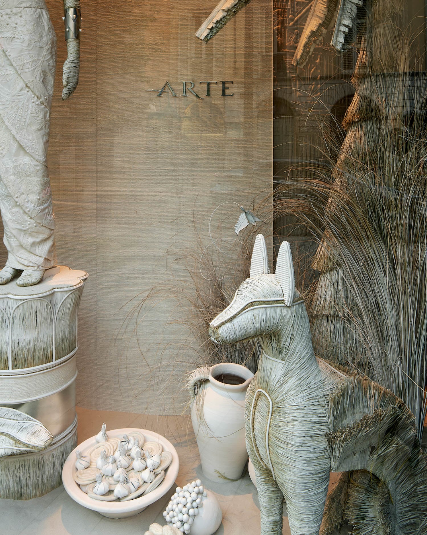 A window display features woven sculptures of a fox, fruit, and vases, along with a partial figure and decorative grass, all in neutral tones. The word "ARTE" is on the background wall.