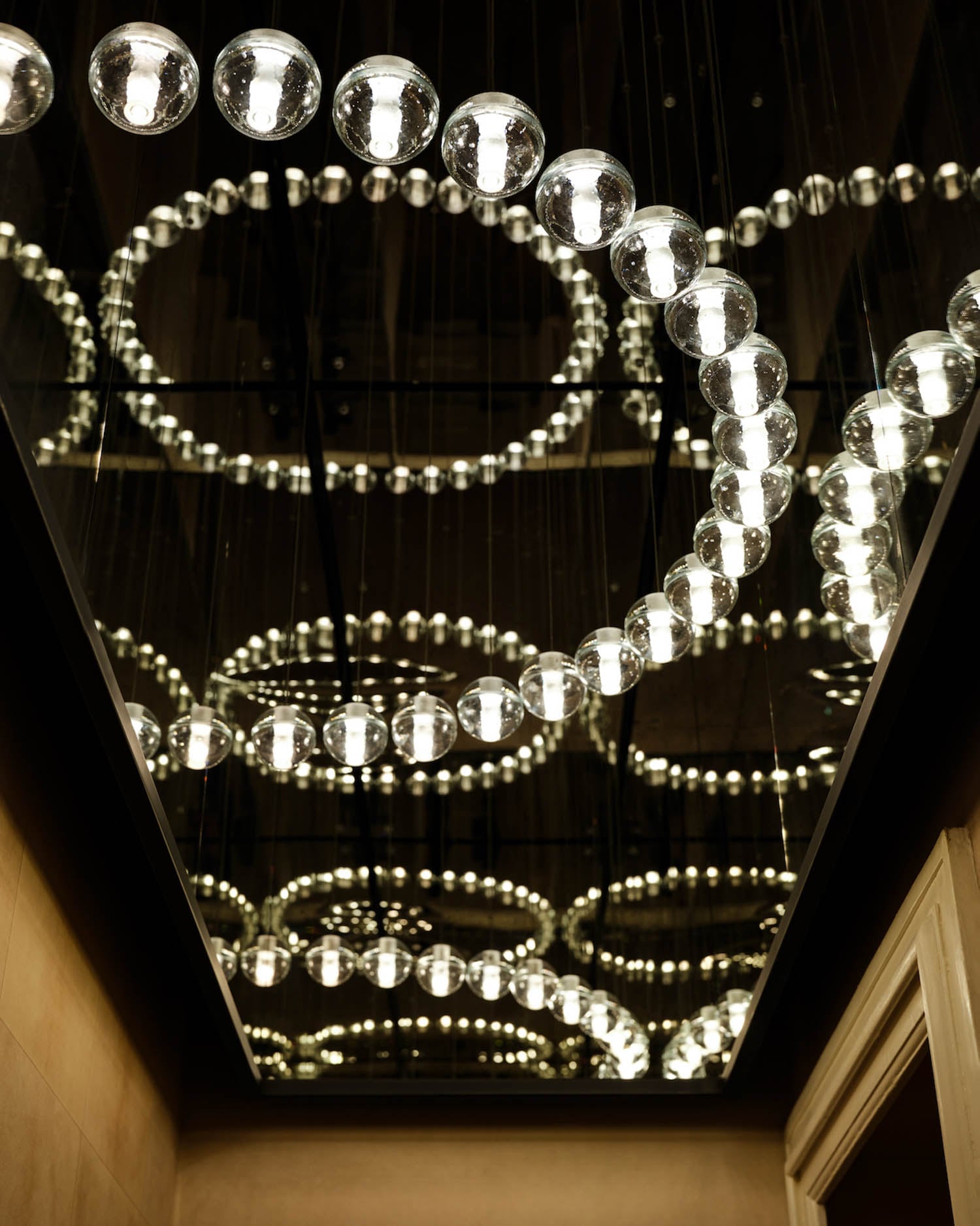 Ceiling installation of spherical light bulbs arranged in circular patterns, reflected in mirrored panels above, creating repeating ring shapes.