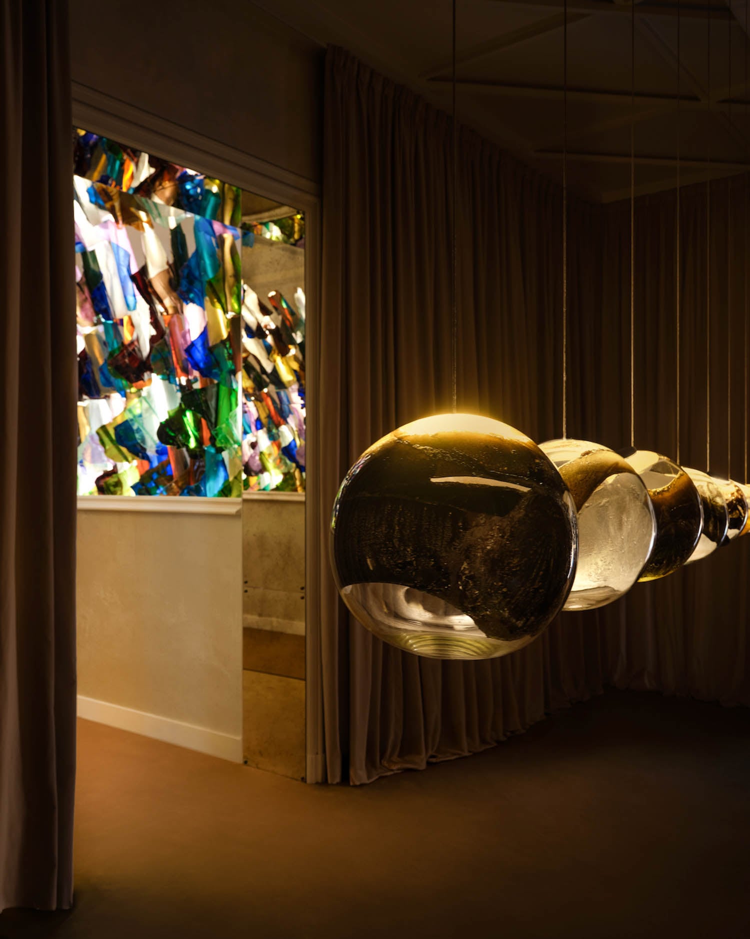 Three large, spherical pendant lights hang in a dim room with beige curtains, near a doorway displaying a colorful glass art installation.
