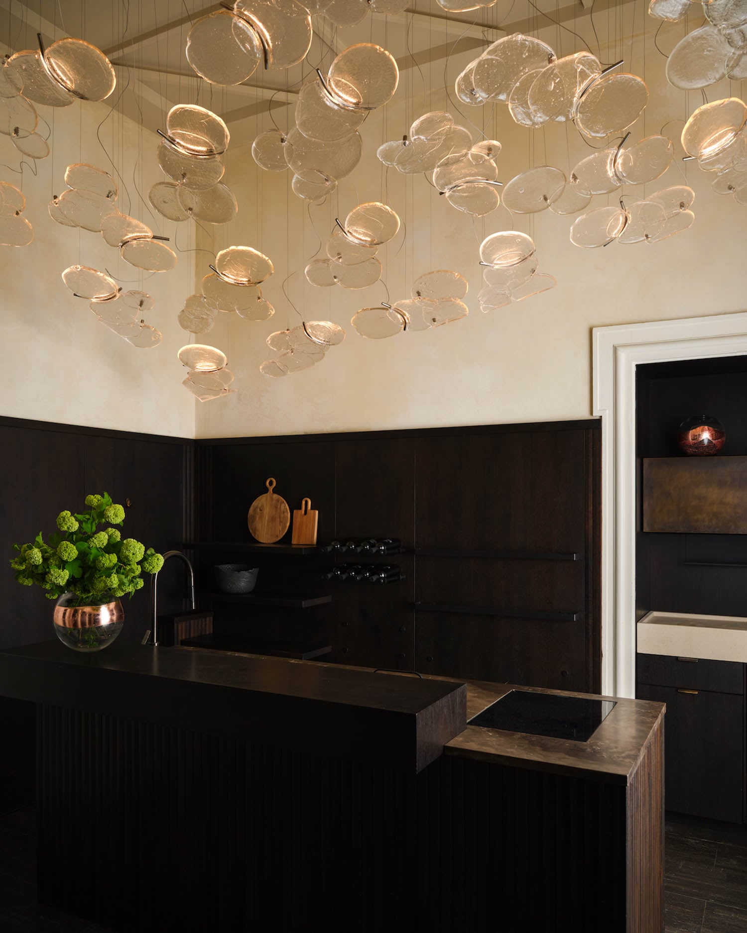 Modern kitchen with dark wood cabinets, minimalist black countertops, a copper vase with green flowers, and a ceiling installation of translucent, disc-shaped pendant lights.