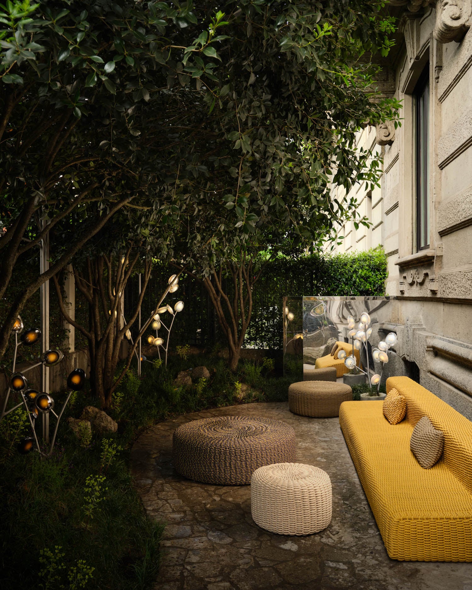 Outdoor patio with wicker tables, a yellow sofa, round poufs, decorative lighting, lush greenery, and a stone path beside a building.