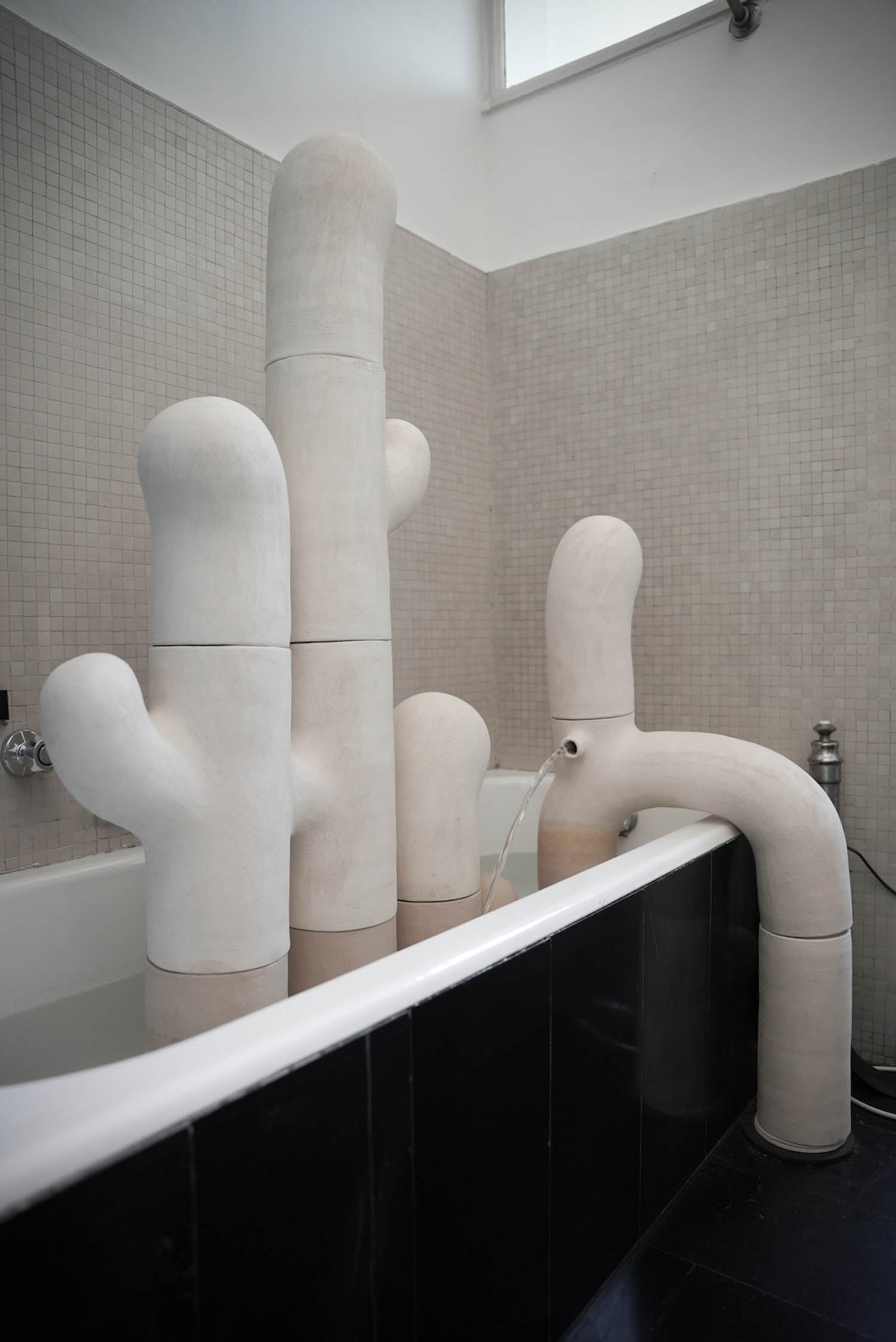 A bathtub filled with sculptural objects resembling abstract, white cacti-like forms, with one feature functioning as a faucet pouring water into the tub.