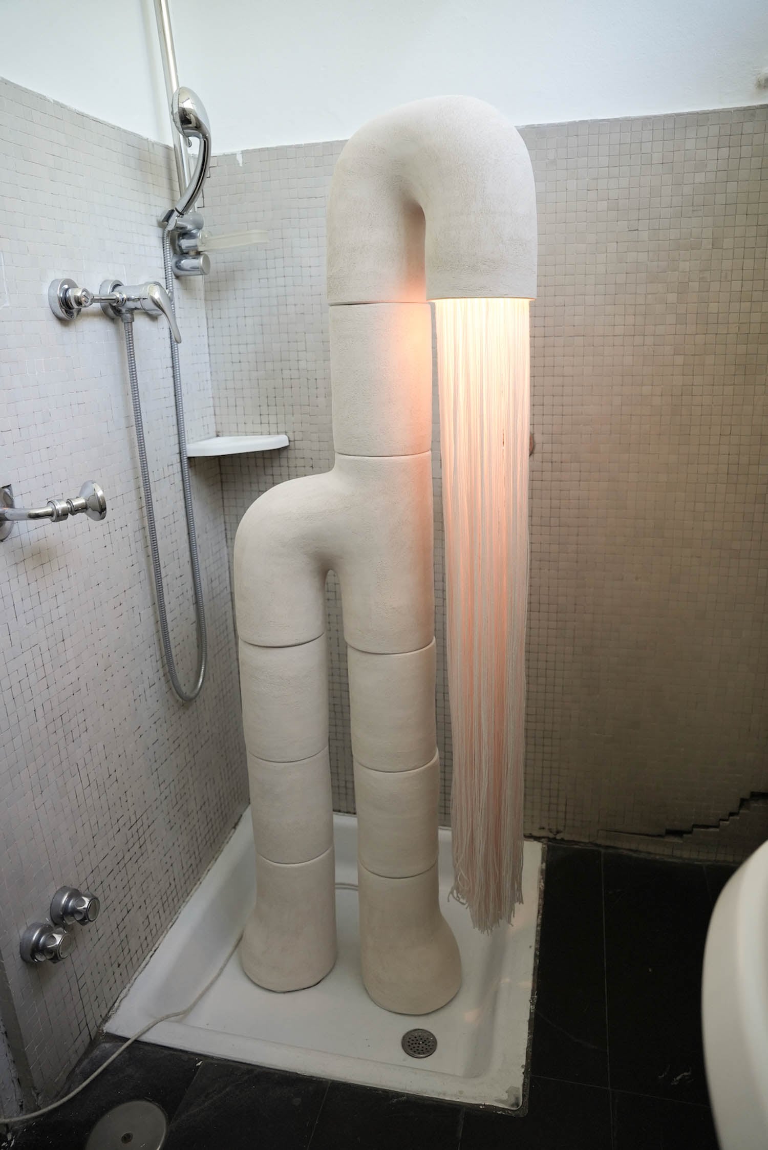 A sculptural lamp shaped like abstract, segmented legs stands in a shower stall; light shines through strands hanging from its top section.