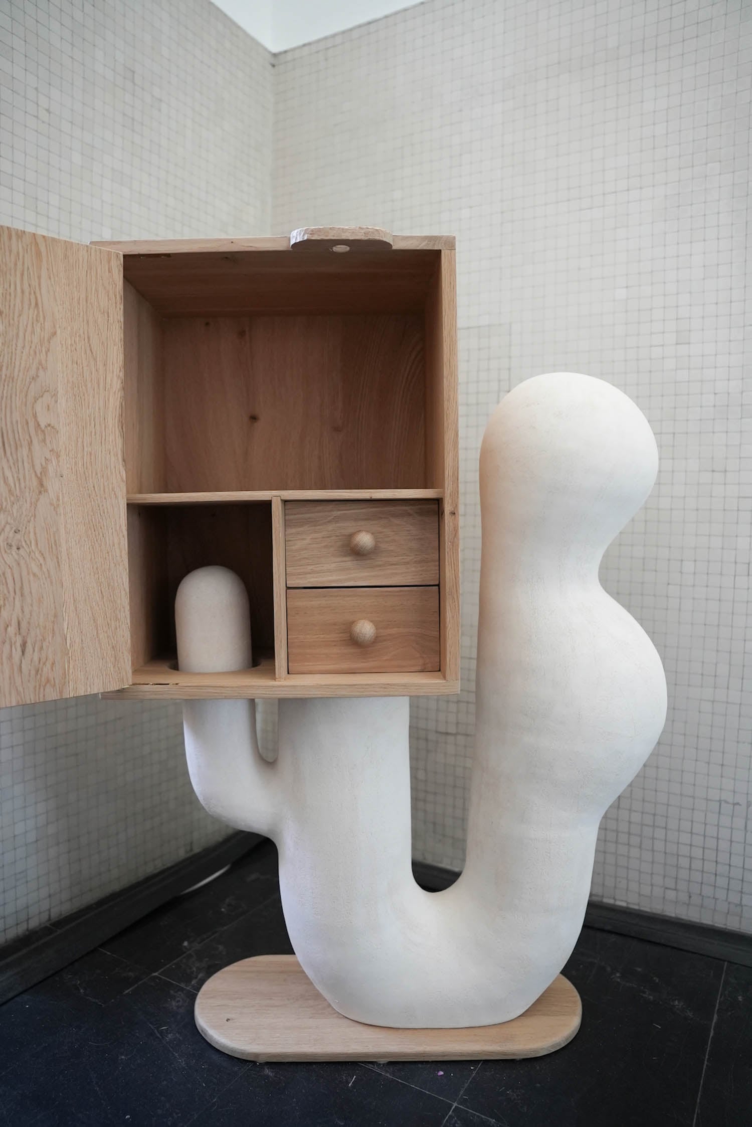 A wooden cabinet with open doors sits atop a white, sculptural base resembling an abstract cactus or organic form, set against a tiled wall.