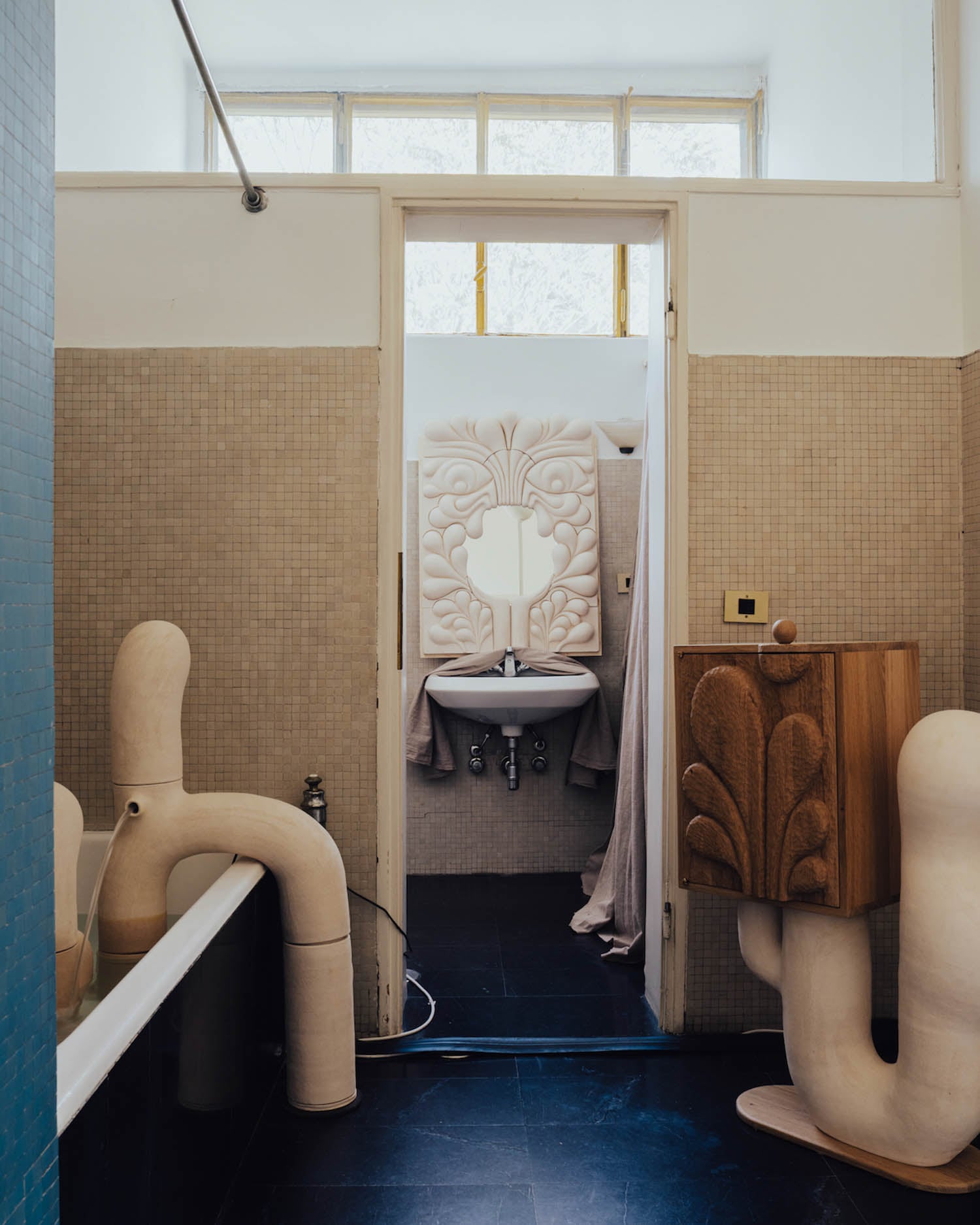 A bathroom with beige tiled walls features an unusual sculptural bathtub and sink, a wooden cabinet with carved designs, and a decorative mirror above the sink.
