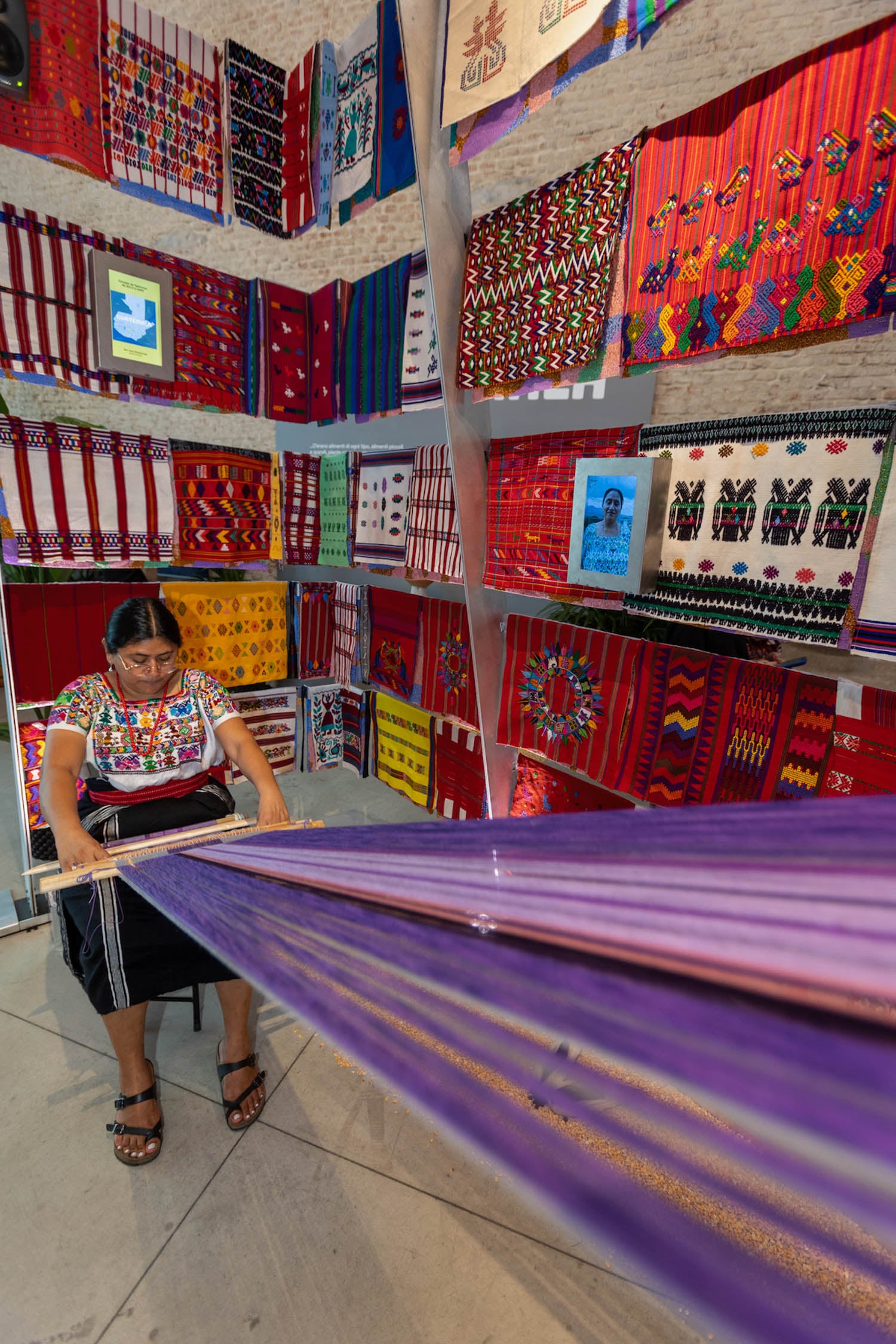 A woman in traditional clothing uses a backstrap loom to weave purple fabric, surrounded by colorful handmade textiles displayed on the walls.