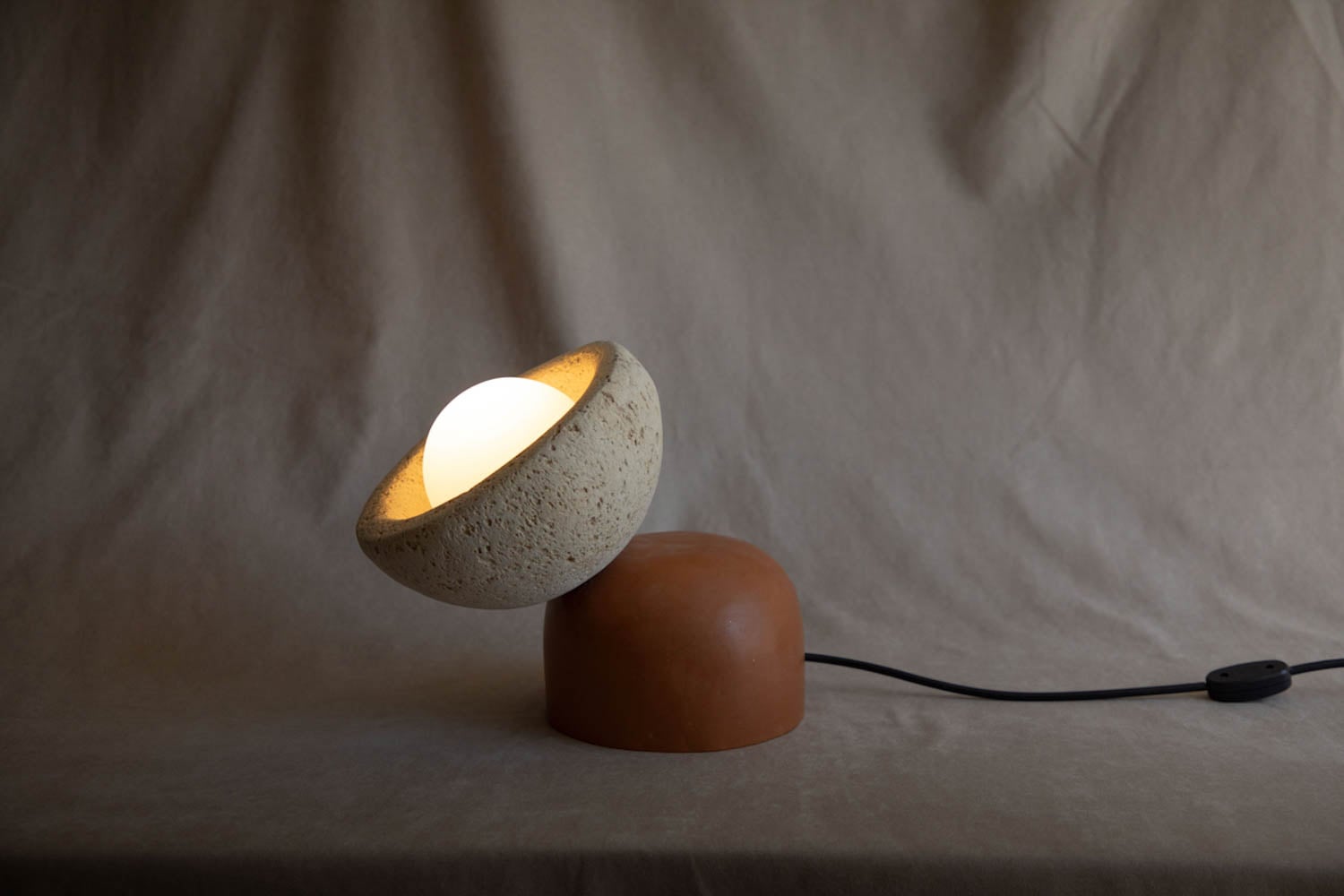 A modern table lamp with a spherical white bulb set in a textured, half-bowl stone shade on a rounded brown base, placed on a neutral fabric surface.