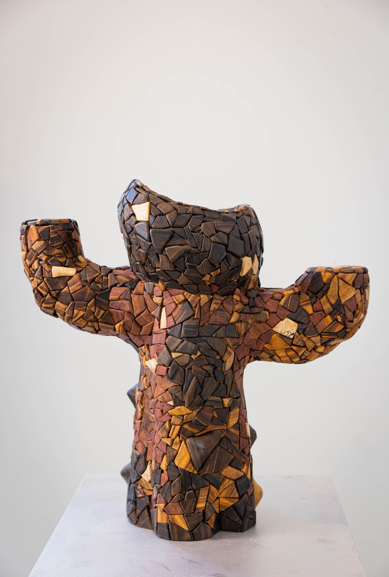 A mosaic sculpture of a bear-like figure made from variously colored wood pieces stands on a white surface against a plain background.