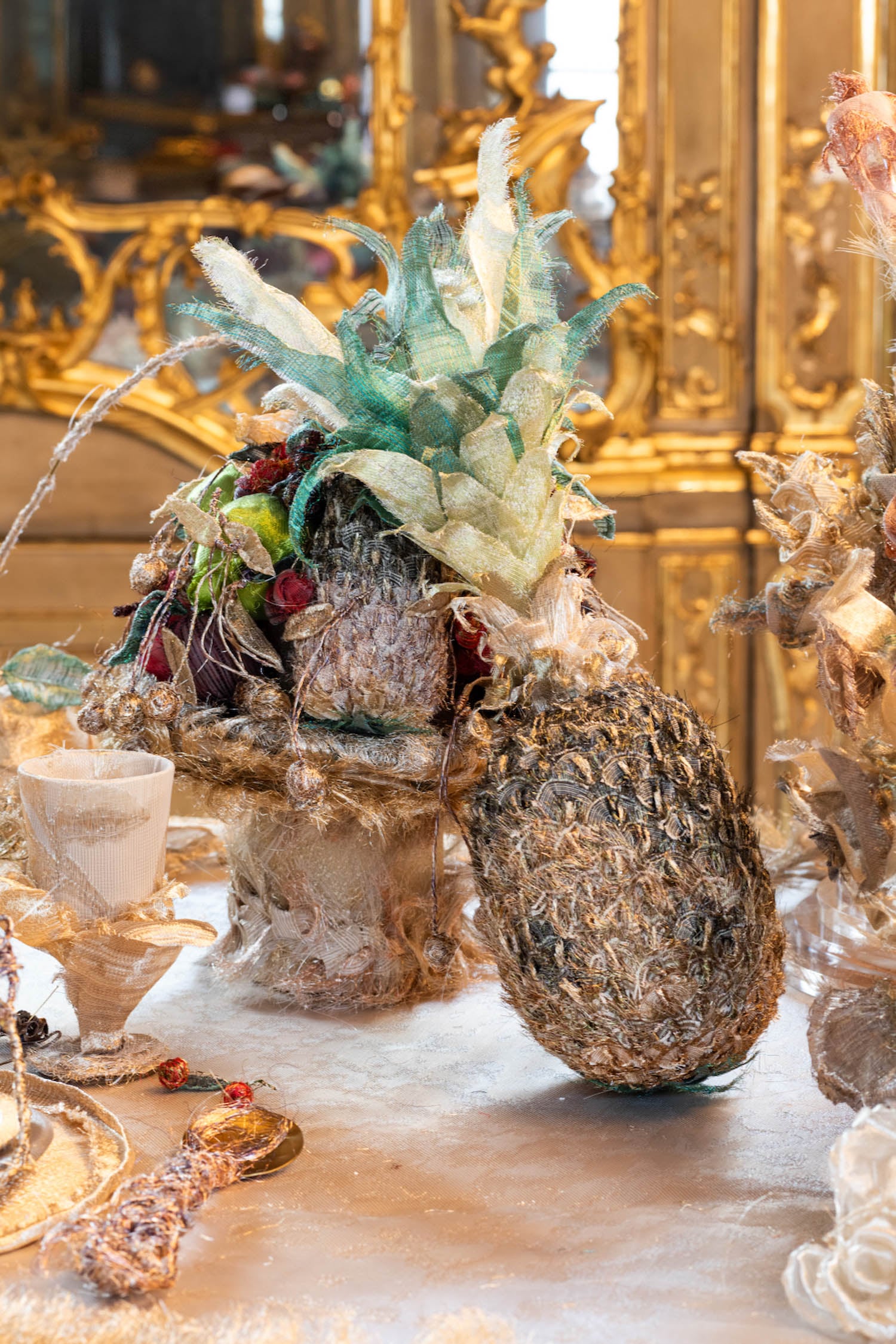 A decorative table setting features a glittering faux pineapple arrangement with gold accents, surrounded by ornate decor and reflective surfaces in a luxurious room.