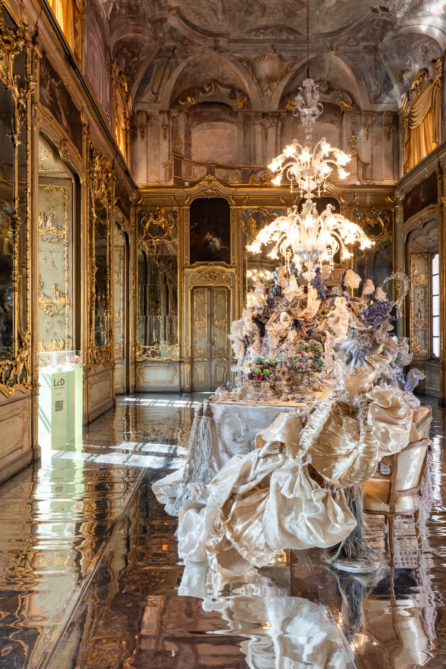 Opulent baroque room with gilded walls, mirrored floors, ornate chandelier, and table set with elaborate decorative sculptures and draped fabric.