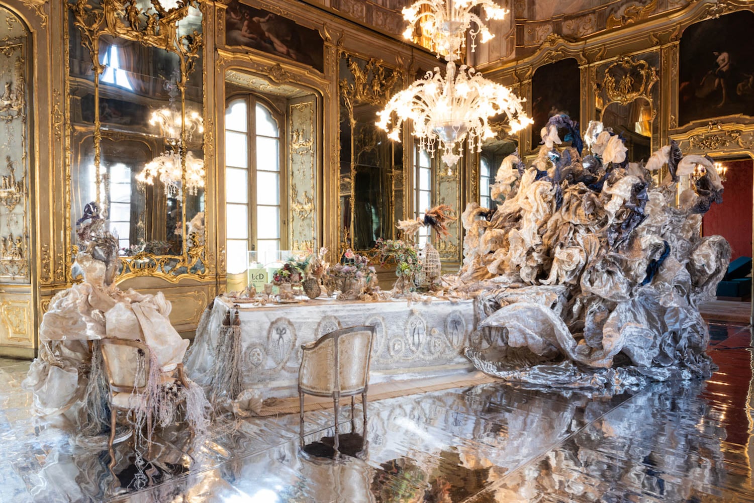Ornate room with gilded walls and mirrors, a chandelier, and a dining table set with elaborate floral arrangements and sculptural decorations, surrounded by translucent, draped materials.