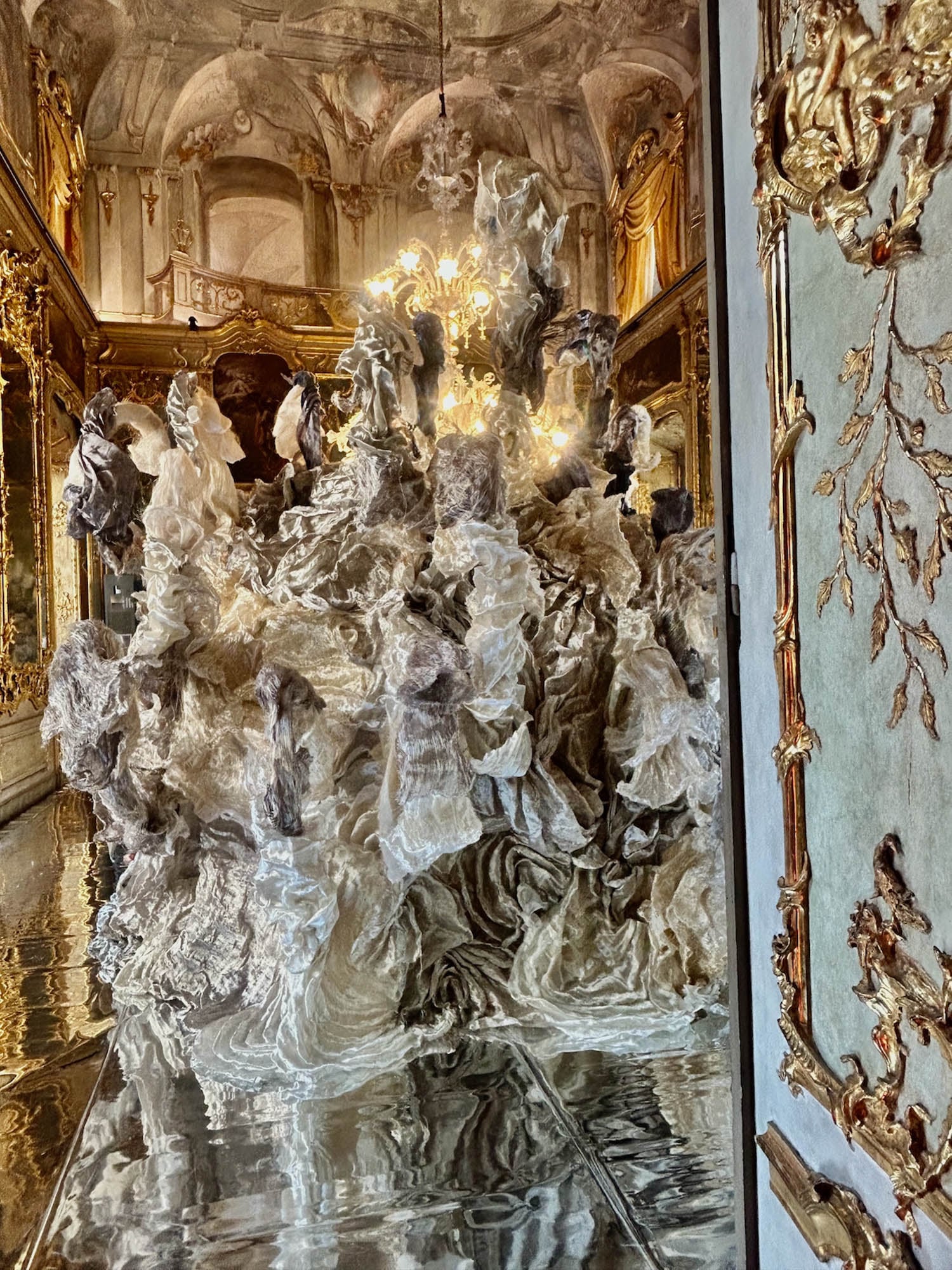 Large abstract sculpture made of white and gray translucent material displayed in an ornate, gold-accented baroque room with a chandelier overhead.