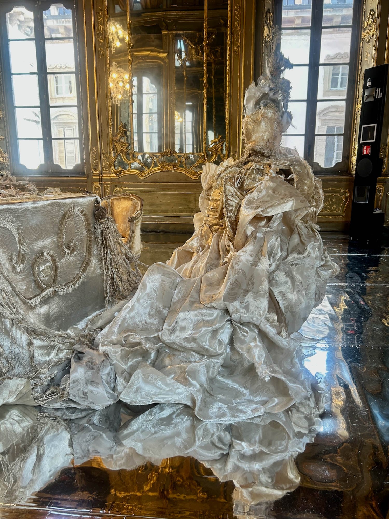 A mannequin in an ornate, historical gown sits in a lavishly decorated room with gold details, mirrored floors, and large windows.