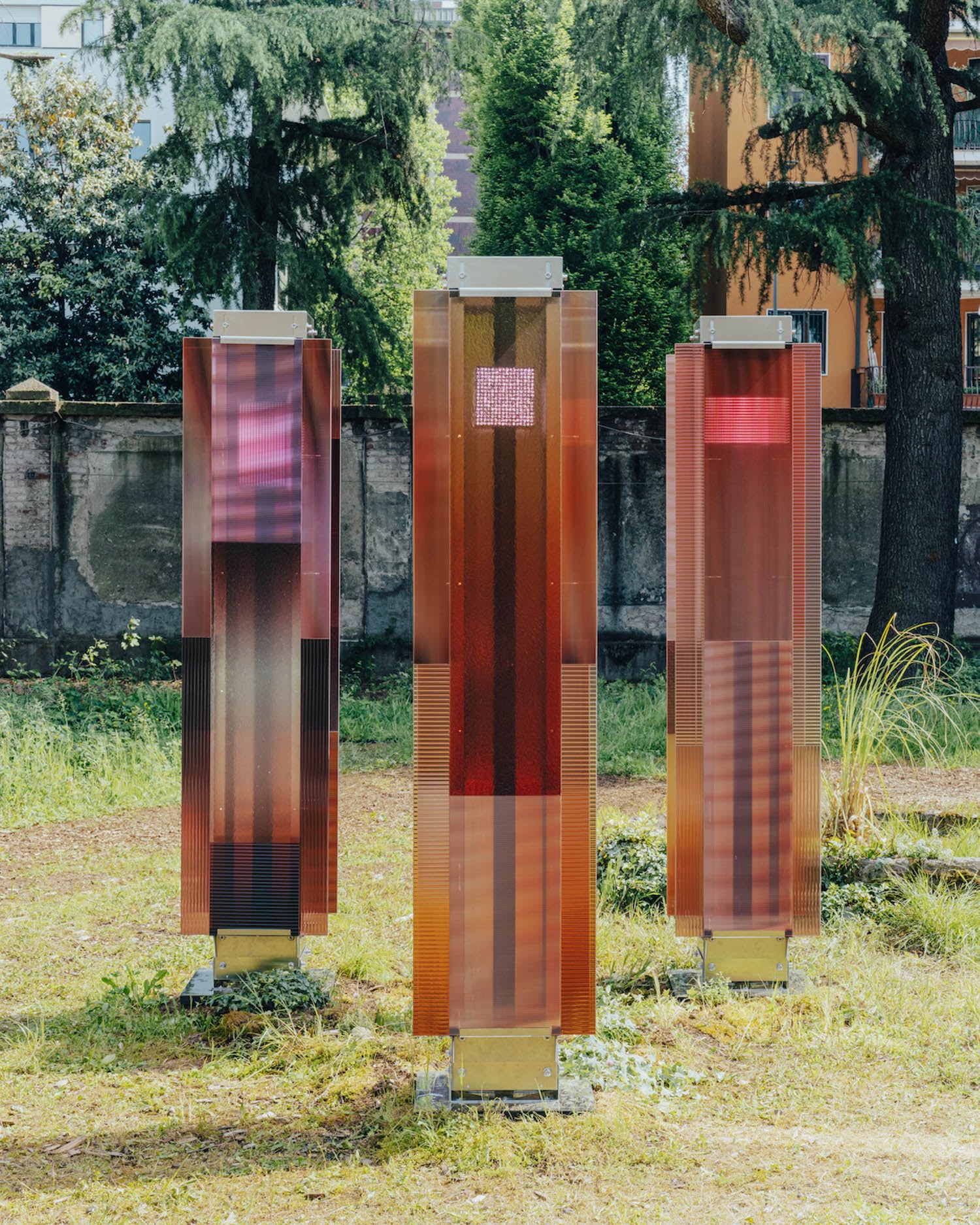 Three vertical, rectangular, translucent red-brown panels stand outdoors on metal bases, surrounded by grass, trees, and nearby buildings.