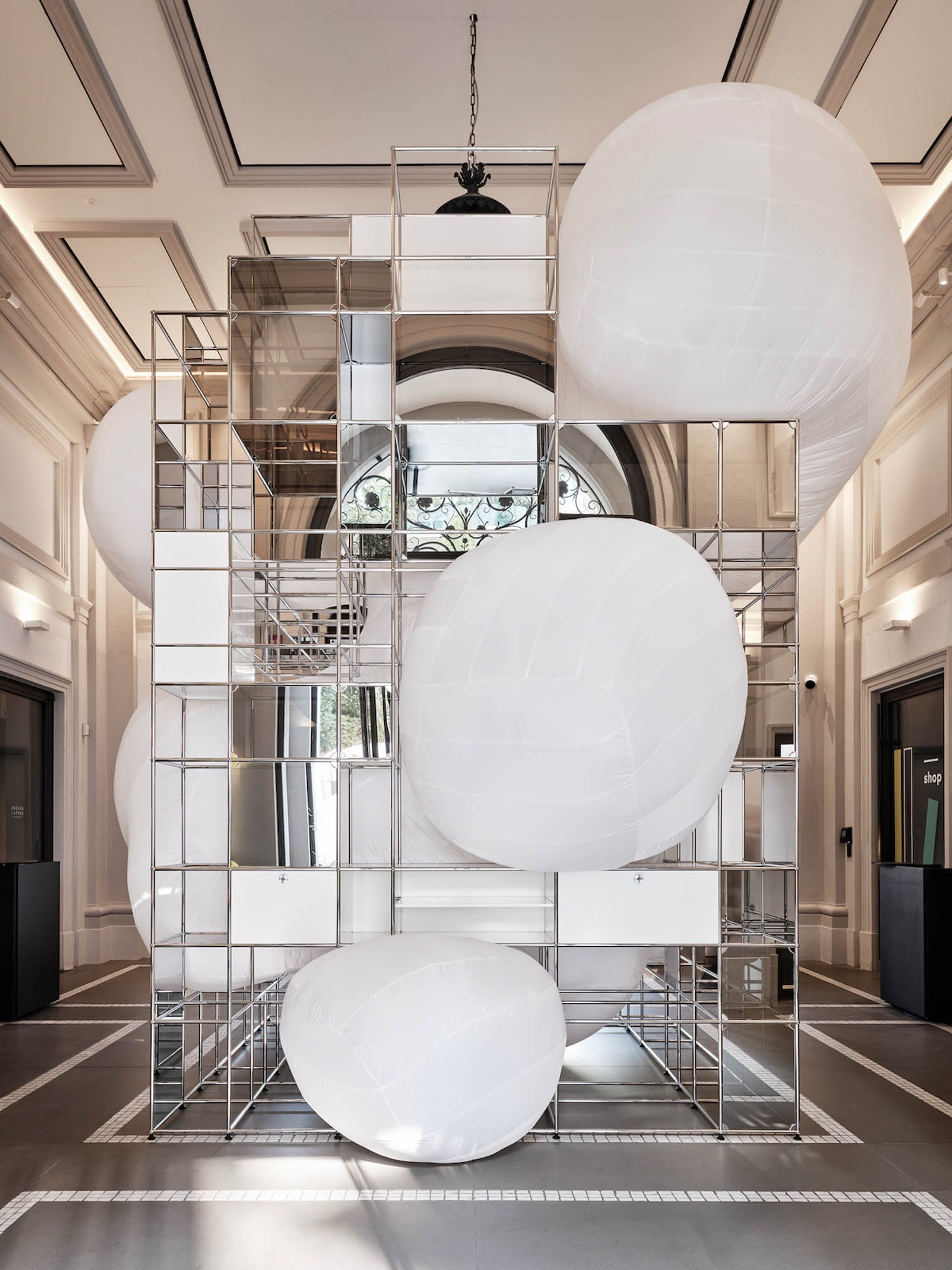 A tall geometric metal structure with large white inflatable spheres is displayed in an elegant, high-ceilinged room with ornate architectural details.