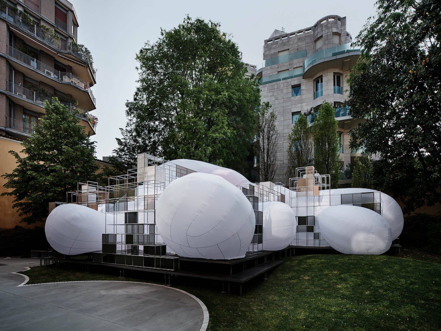 A contemporary outdoor art installation featuring large white inflated shapes attached to a grid-like metal structure, set on a grassy area with modern buildings and trees in the background.