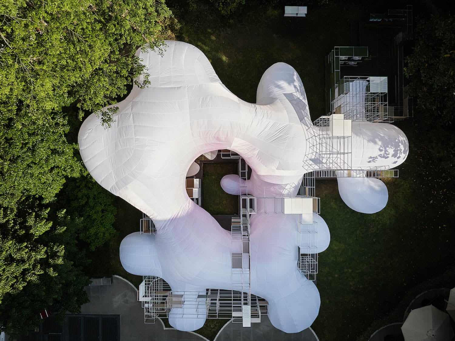 Aerial view of a large, white, organic-shaped inflatable structure with interconnected sections, set on green grass and surrounded by trees.