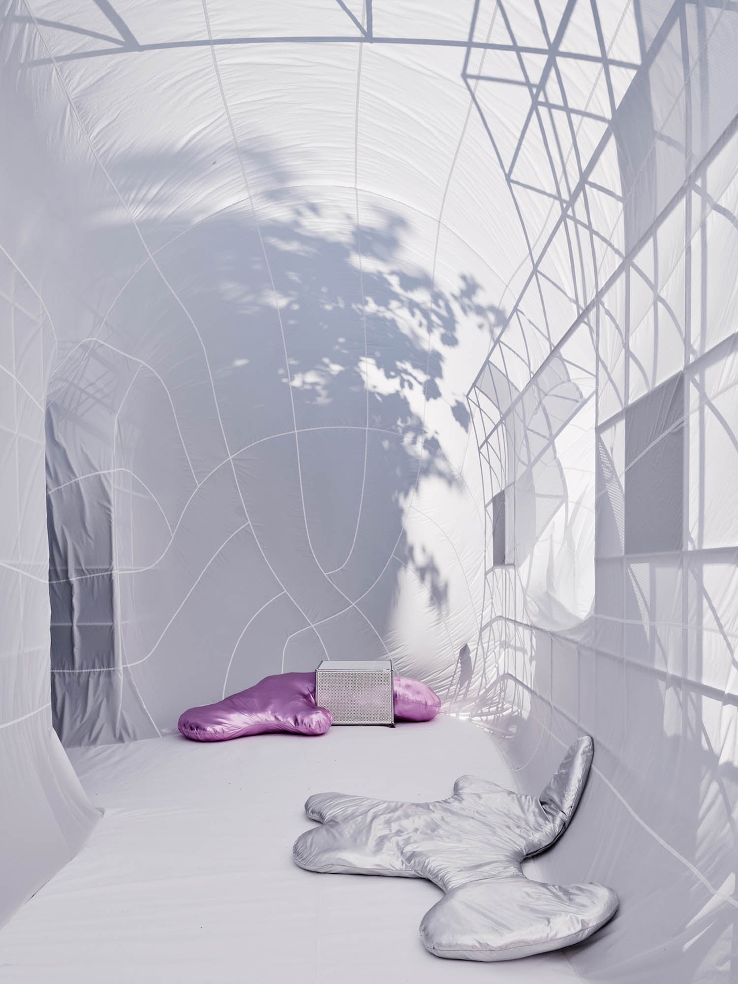 A white, tent-like room with web-like seams, featuring abstract silver and purple cushions and a small white radiator, lit by natural light and shadows of tree branches.