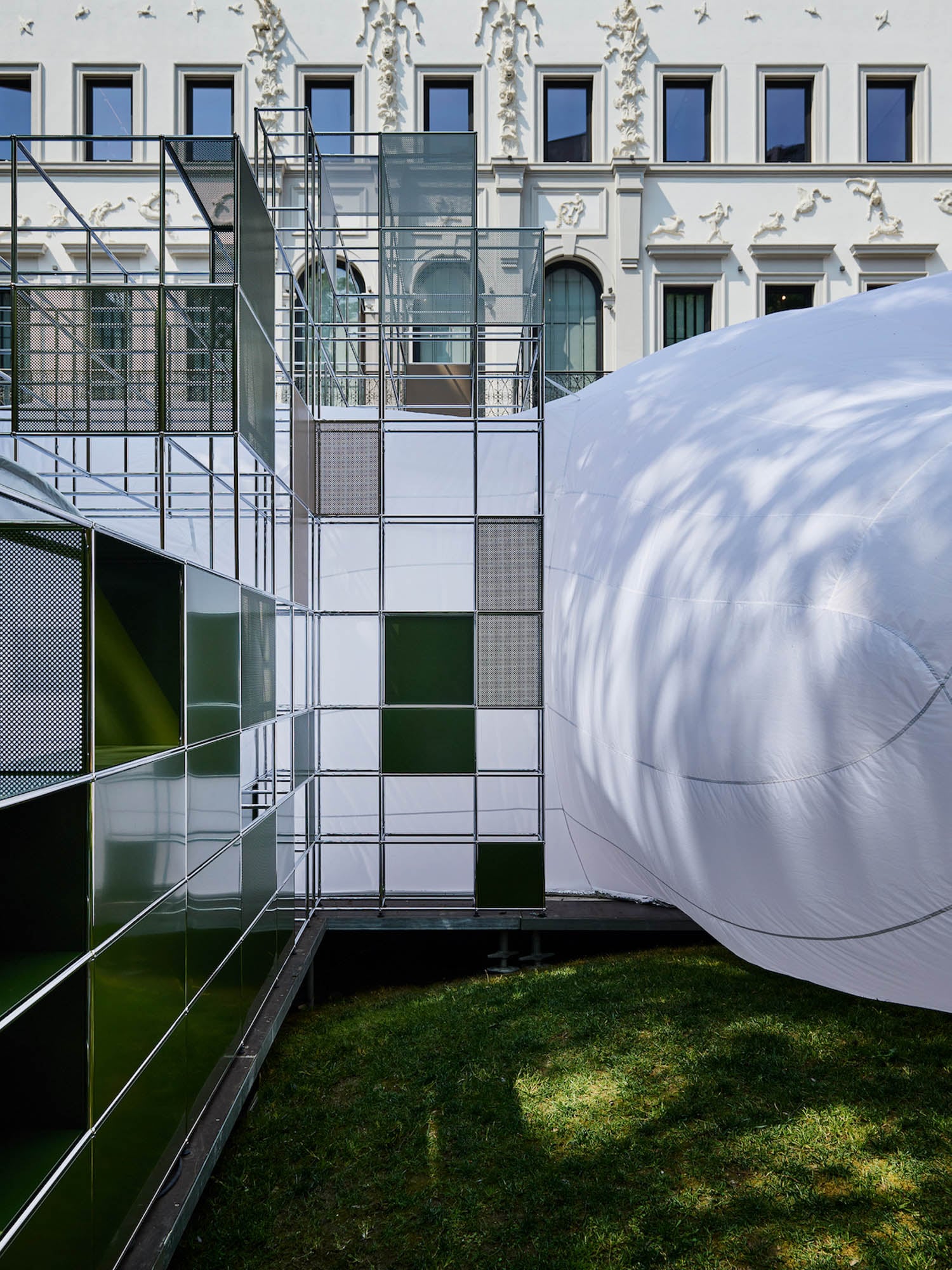 A modern outdoor installation features a metal grid structure with green and white panels next to a large, inflated white form in front of a decorative white building.