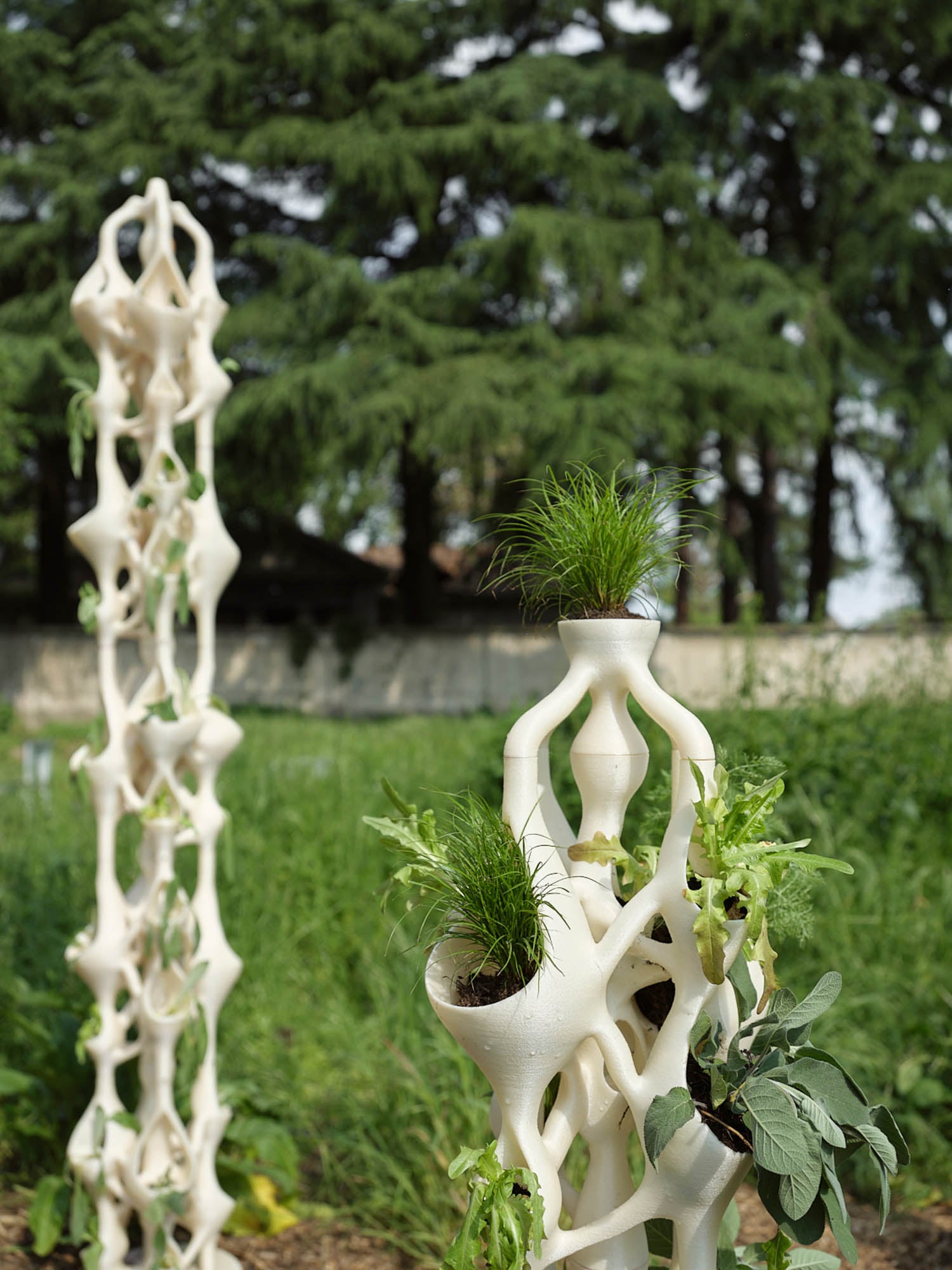 3D-printed white plant structures with various green plants growing in them are set outdoors in a grassy area with trees in the background.