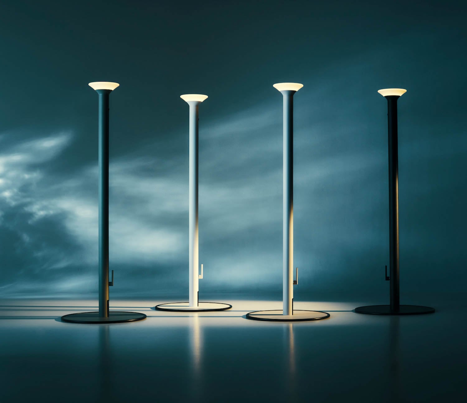 Four modern floor lamps, three white and one black, stand upright on a smooth surface with a blue gradient background and soft shadows.