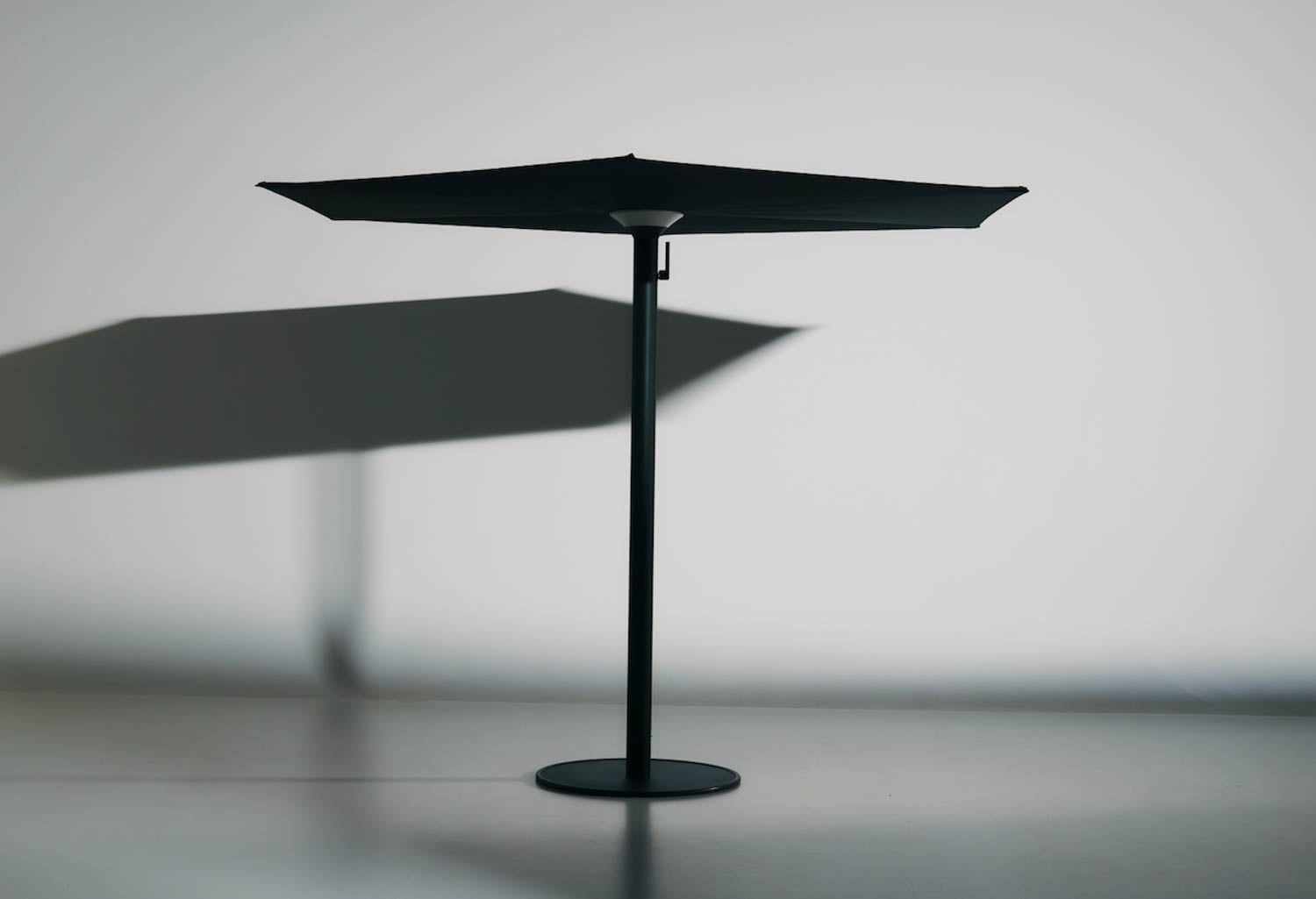 A black patio umbrella with a round base stands on a smooth floor, casting a shadow on a plain white wall behind it.