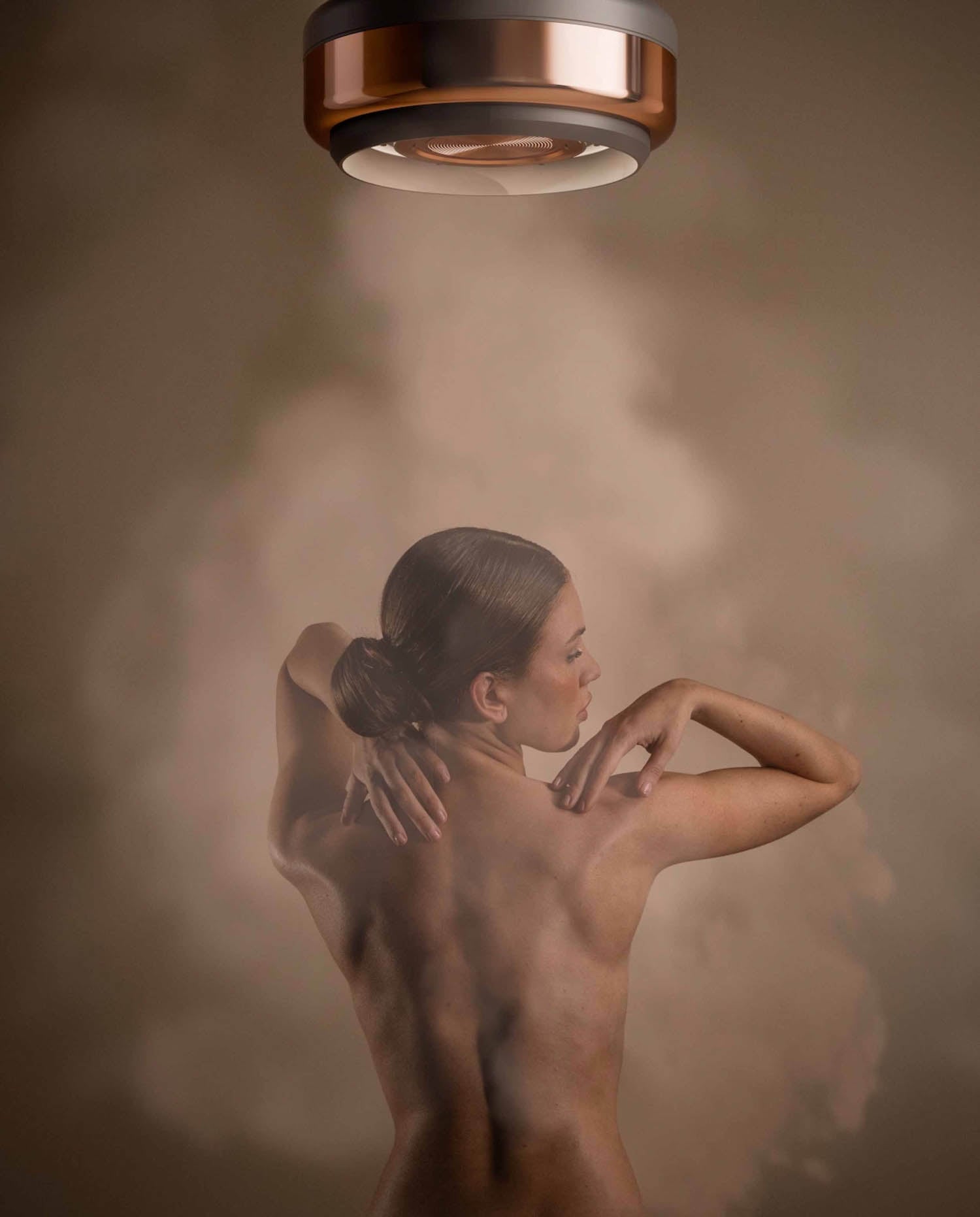 A woman with her back to the camera stands under a circular ceiling device emitting steam or mist, with her arms raised and touching her shoulders.