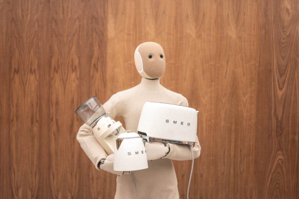 A humanoid figure holding three white SMEG kitchen appliances in front of a wooden wall.