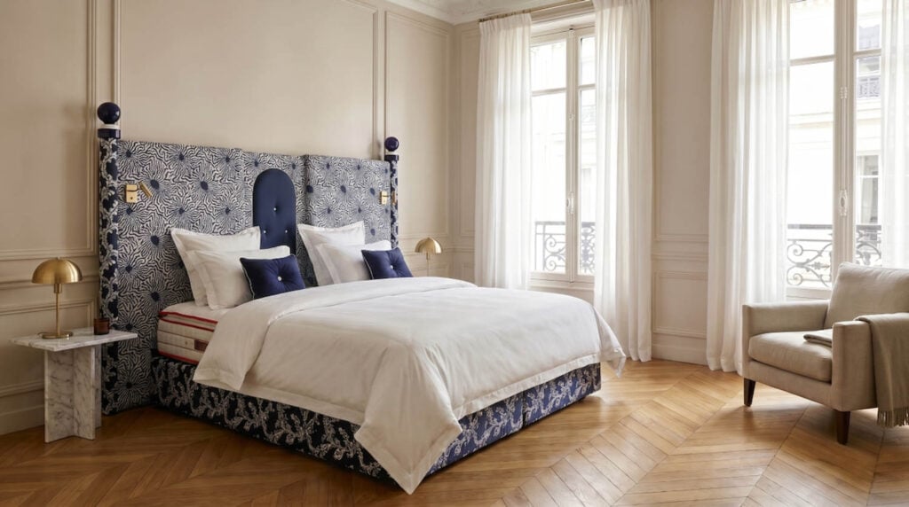 A neatly made bed with a patterned headboard, two bedside tables with lamps, large windows with sheer curtains, and a beige armchair in a bright, elegant bedroom.