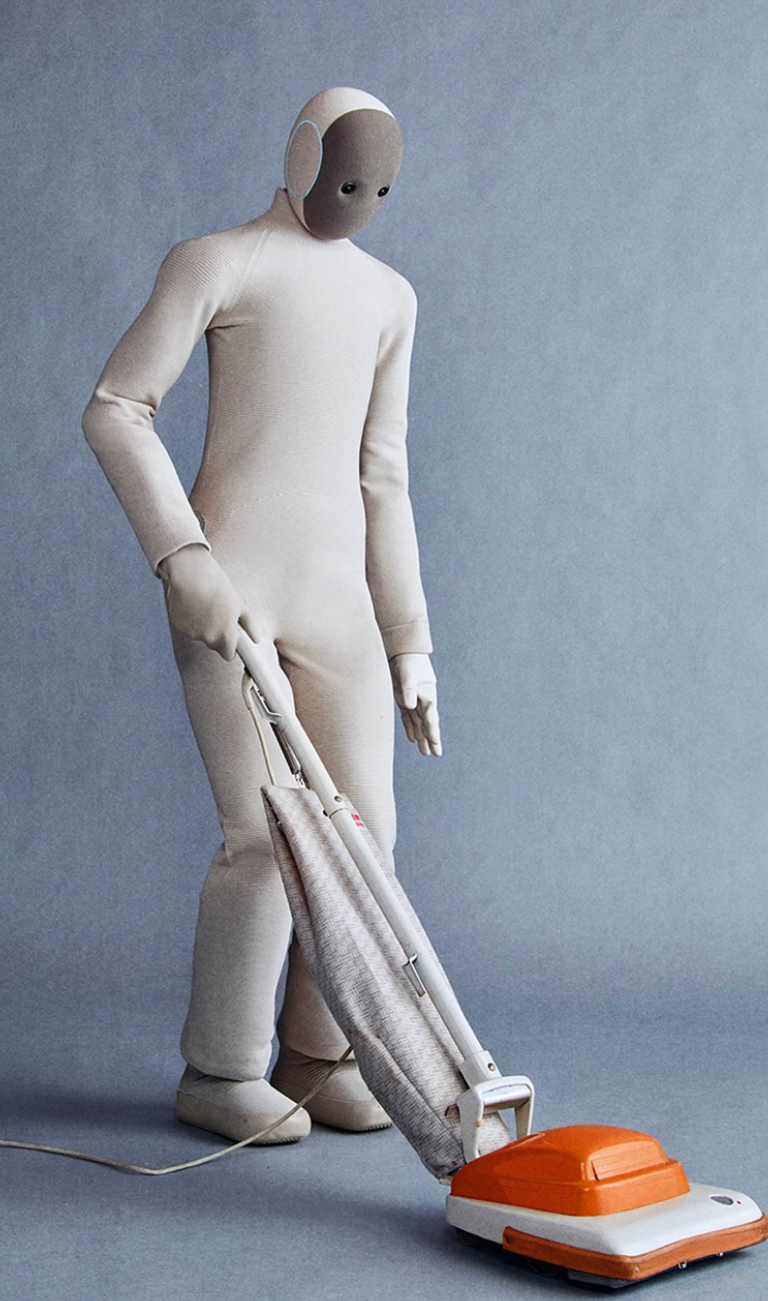 A humanoid figure in a beige bodysuit vacuuming the floor with an orange and white vacuum cleaner against a plain gray background.