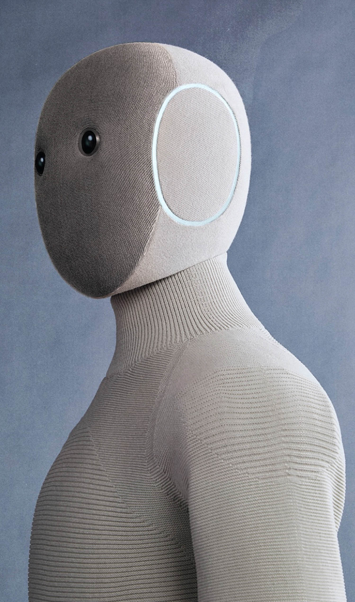 A side view of a humanoid robot with a textured, fabric-like surface, round black eyes, and a circular detail on the side of its head, against a plain gray background.