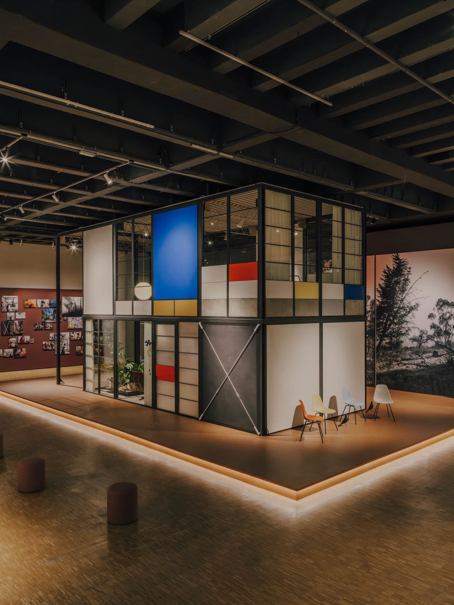 A modern, box-shaped structure with glass walls and geometric colored panels sits in a spacious, well-lit gallery with photos on the walls and a few chairs arranged nearby.