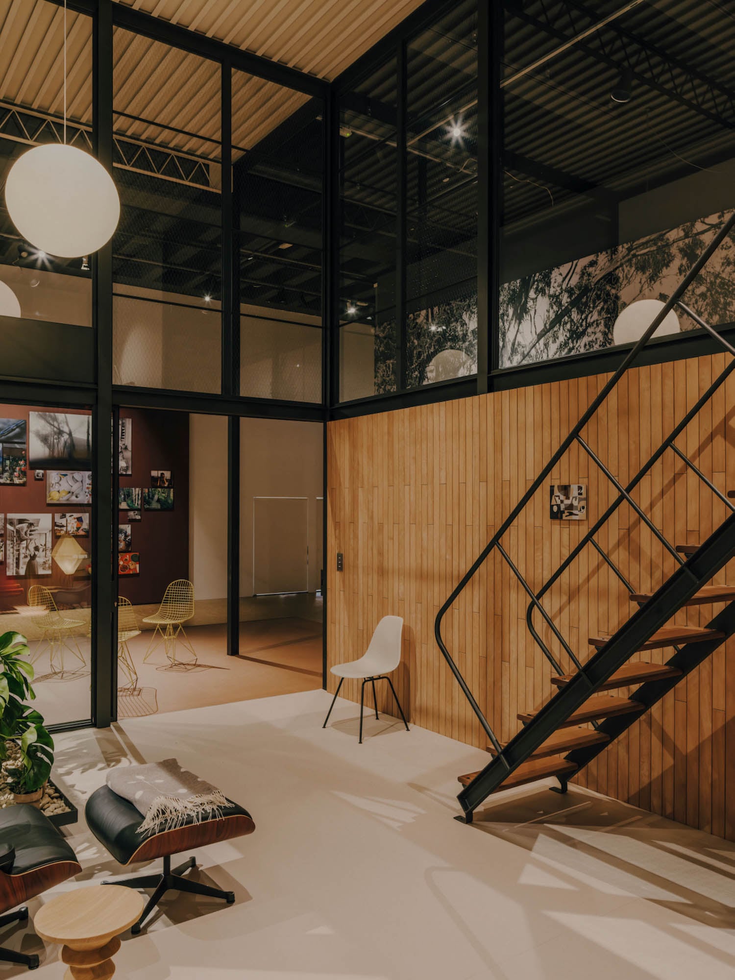 Modern loft interior with wooden walls, a black metal staircase, glass-paneled upper level, and minimal furniture including a white chair, lounge seat, and indoor plants.