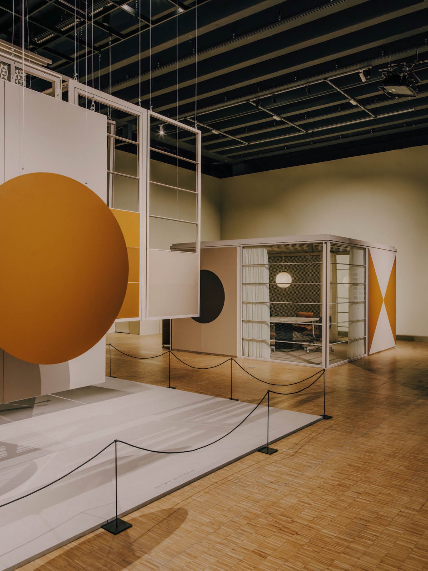 Exhibition space with modern geometric art installations featuring large orange and black circles and an enclosed area with transparent walls, on a wooden floor with black stanchions.
