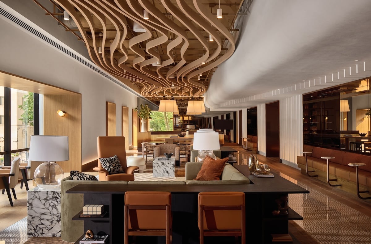 Modern hotel lobby with contemporary furniture, marble tables, decorative lamps, and a wavy wooden ceiling feature—design elements inspired by the NYCxDESIGN Awards. Large windows bring in natural light.