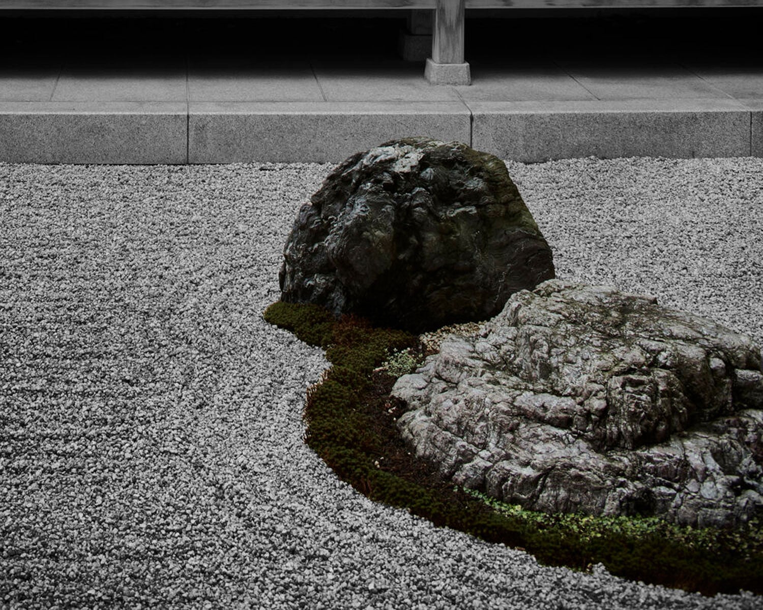 Two large rocks surrounded by moss and raked gravel in a minimalist Japanese Zen garden, with a stone step and wooden structure in the background.