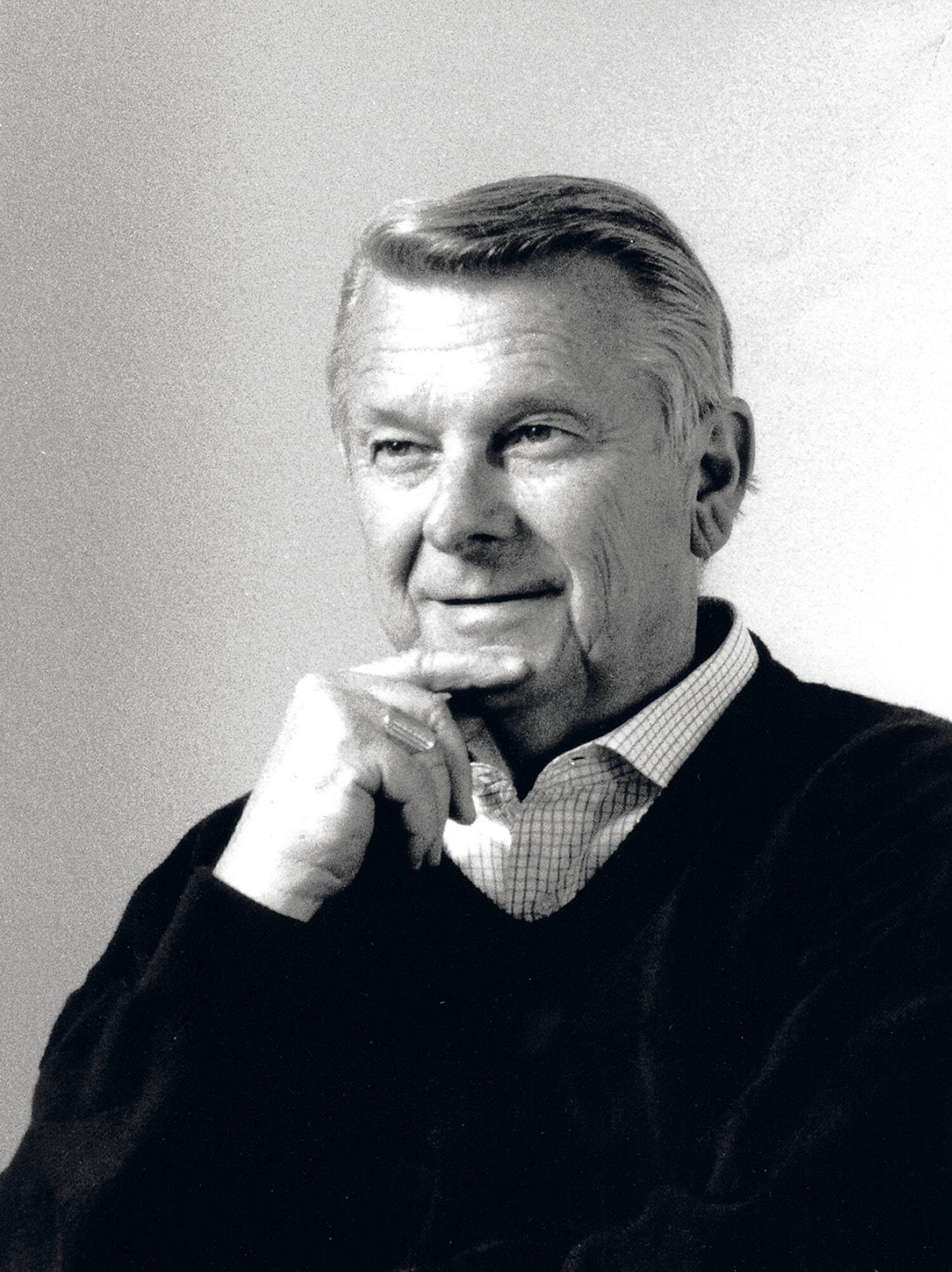 Older man with light hair and a thoughtful expression, wearing a collared shirt and sweater, resting his chin on his hand against a plain background.