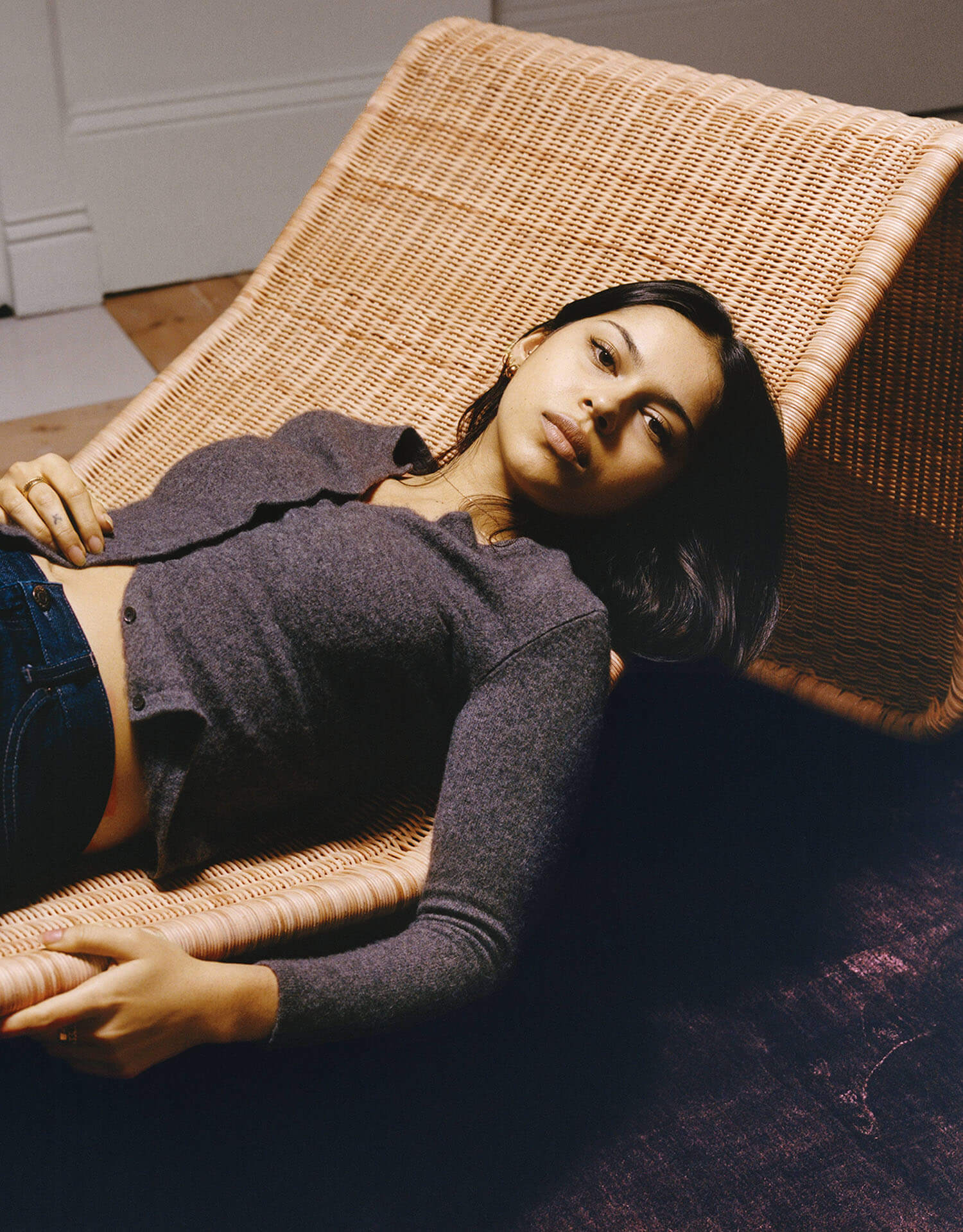 A person with long dark hair lies on a woven lounge chair, wearing a gray cardigan and jeans, looking toward the camera in a softly lit room.